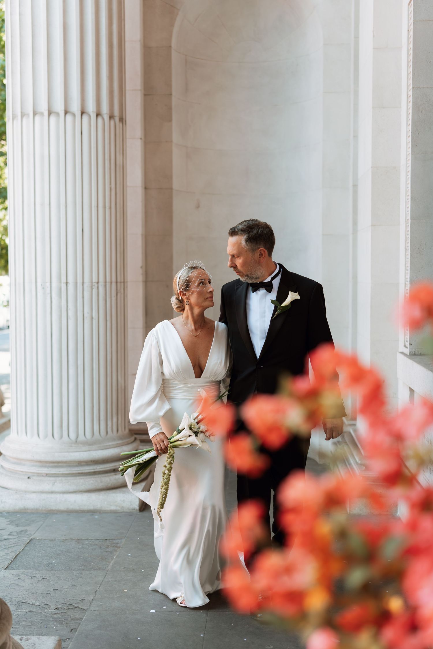 A couple in wedding attire share an intimate moment between elegant white columns with coral flowers in the foreground.