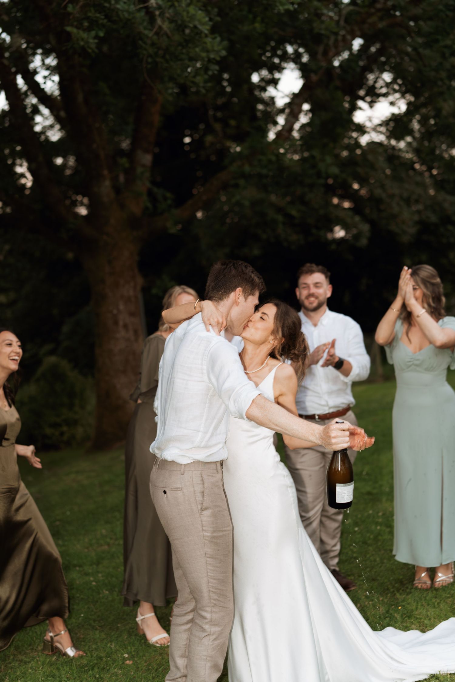 Wedding party dancing and celebrating outdoors on green lawn with natural backdrop.