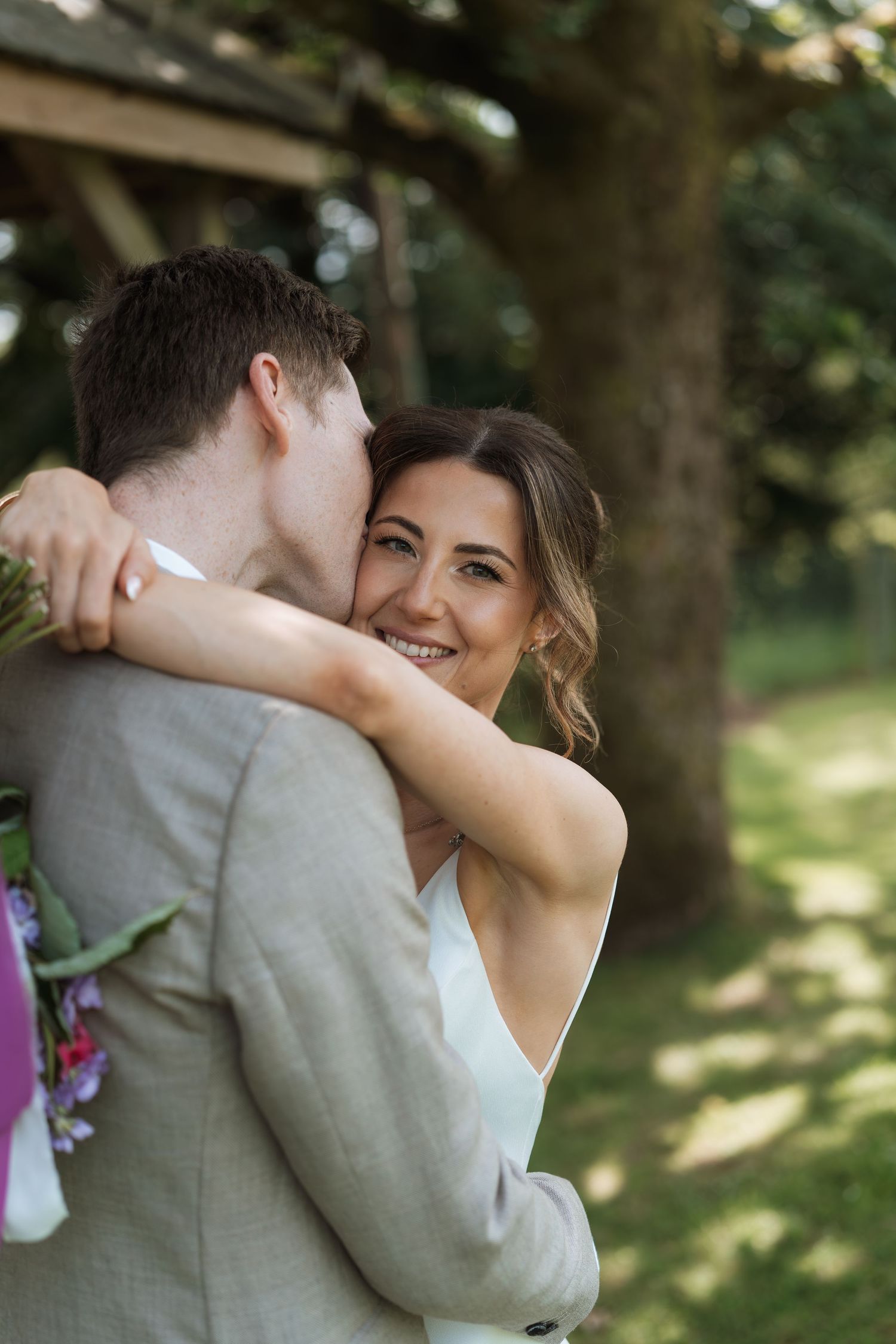 A romantic outdoor wedding moment captured between a couple embracing under trees in natural lighting.