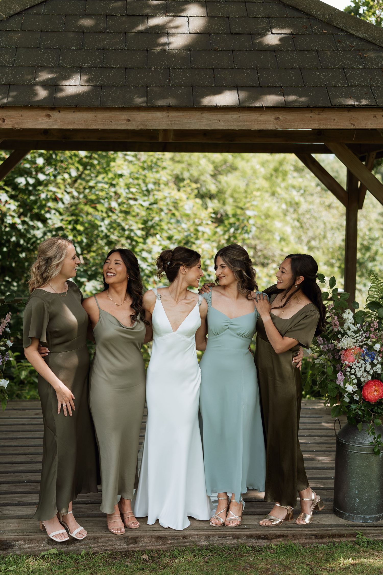 Wedding party in coordinated dresses posing under a covered outdoor pavilion.