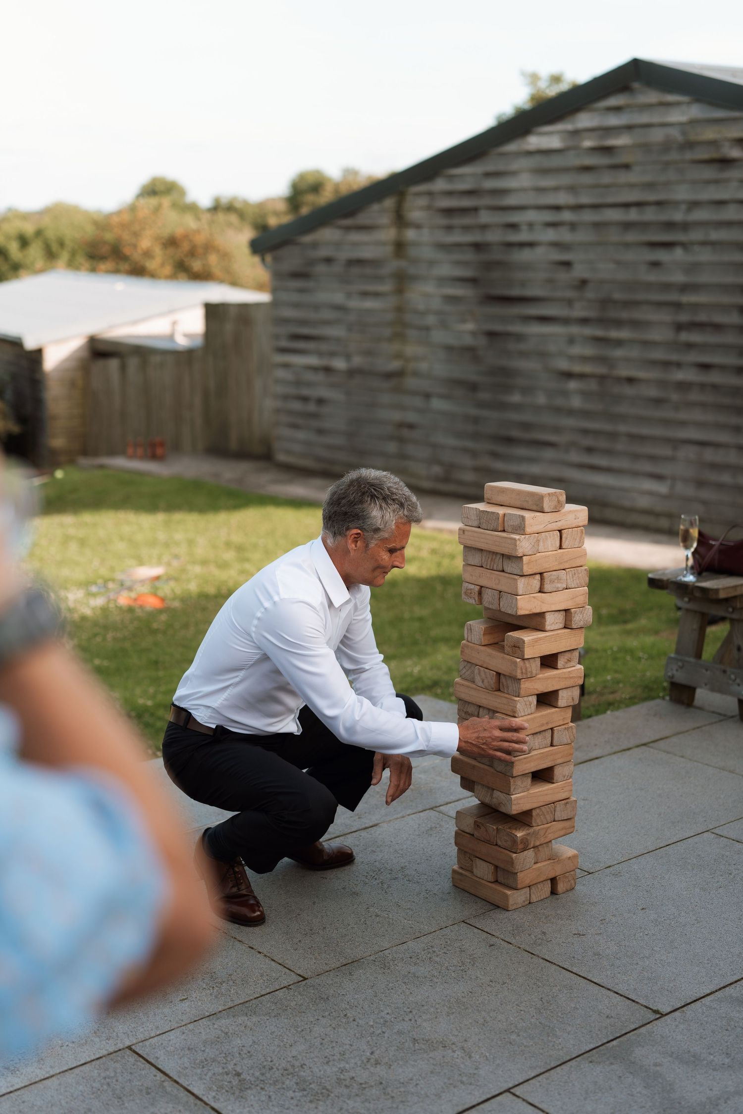 A person crouches down to examine a large Jenga tower set up on an outdoor patio.