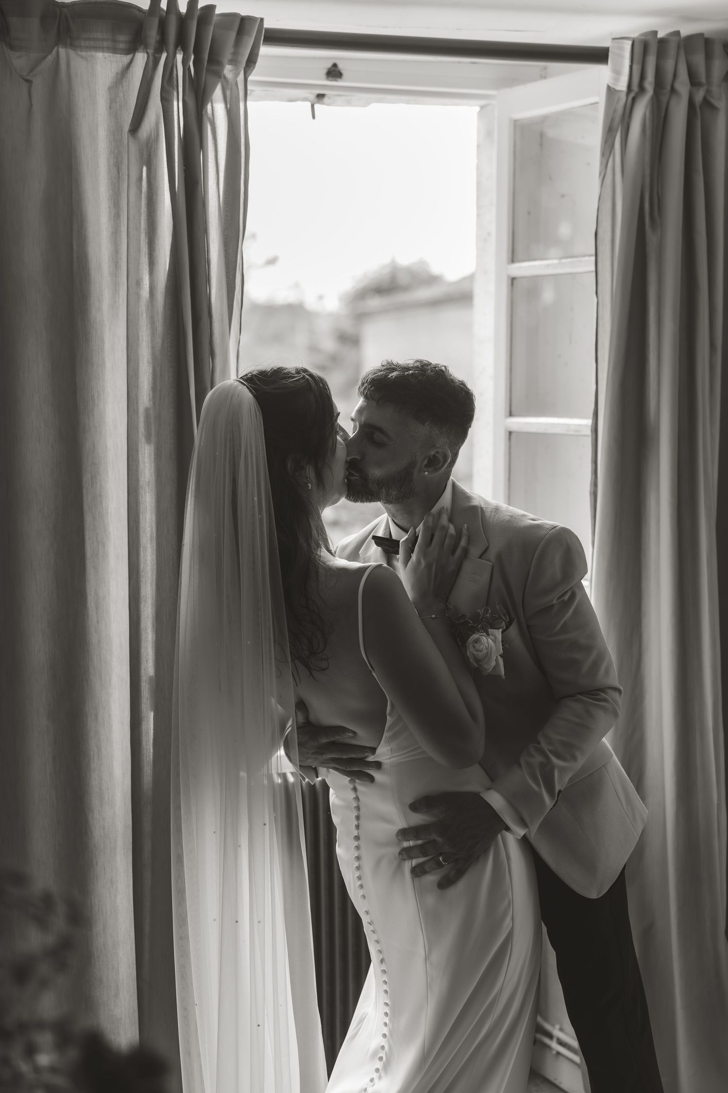 Intimate black and white photo of couple sharing tender moment by window with curtains.