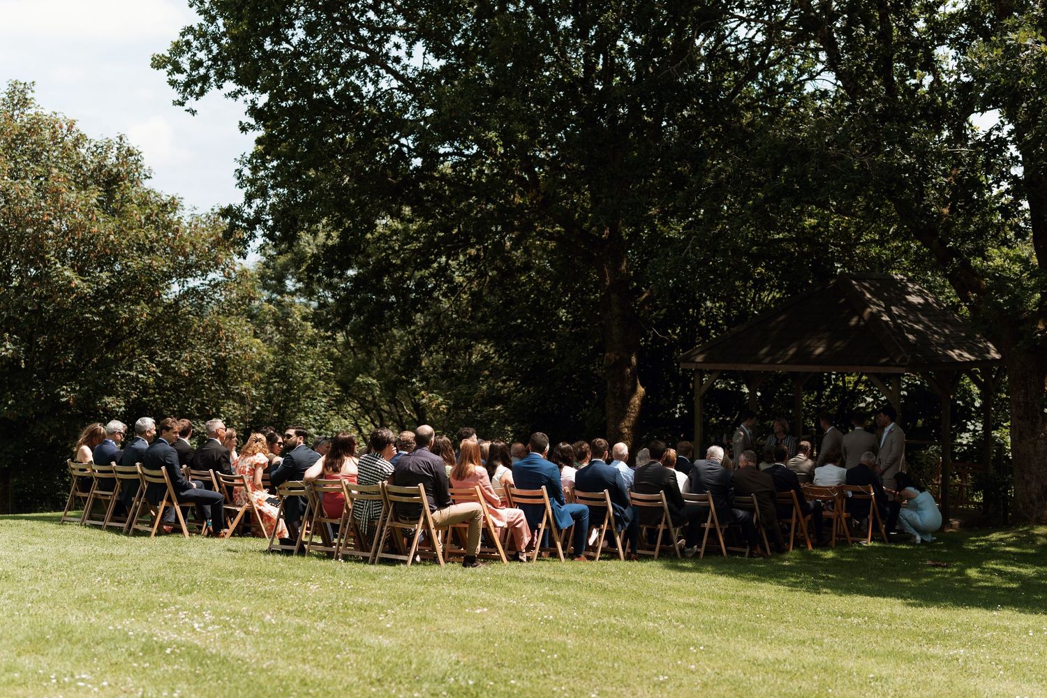 Wide shot of outdoor wedding ceremony with guests seated under trees.
