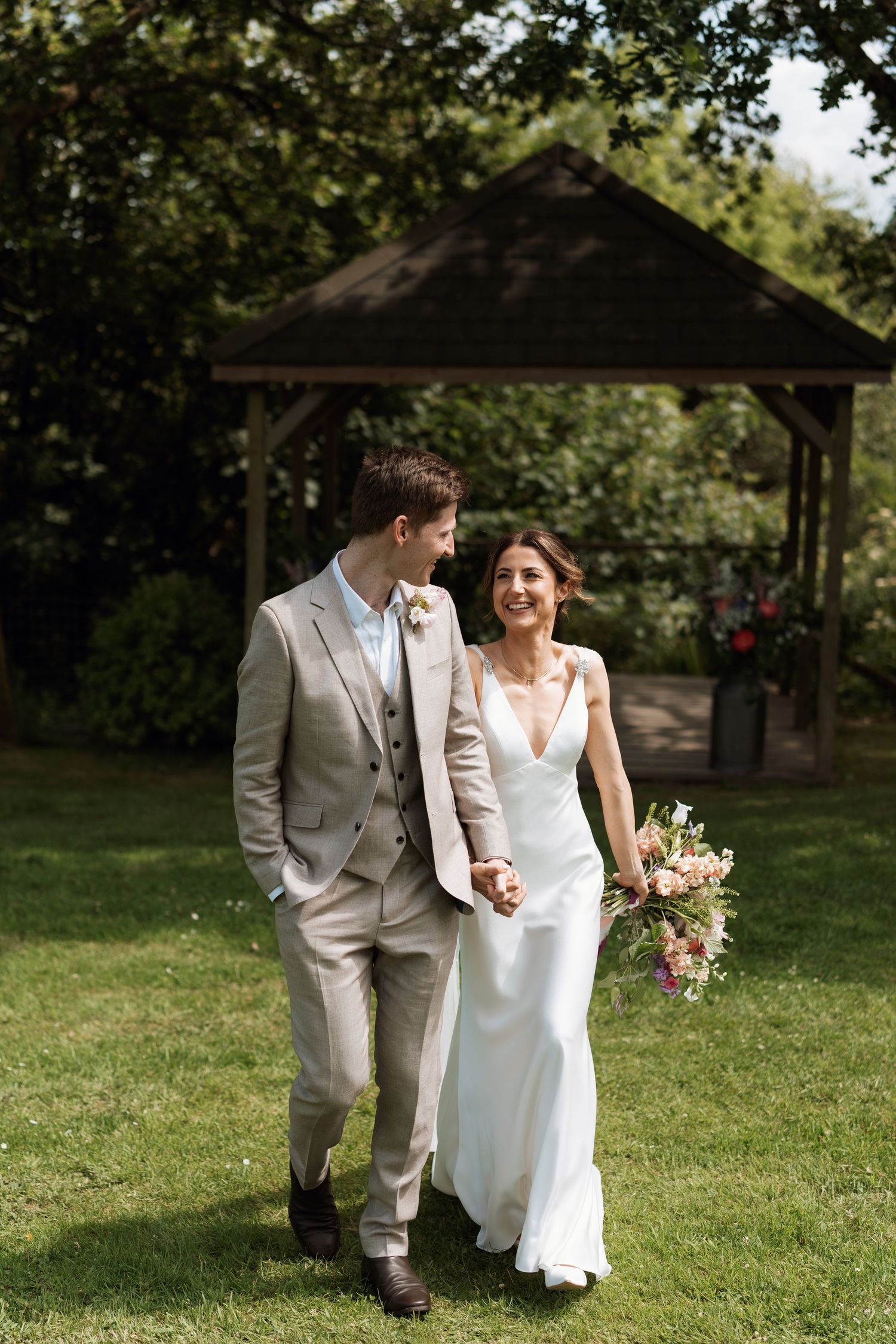 Couple walking hand in hand through a garden with rustic gazebo in background.