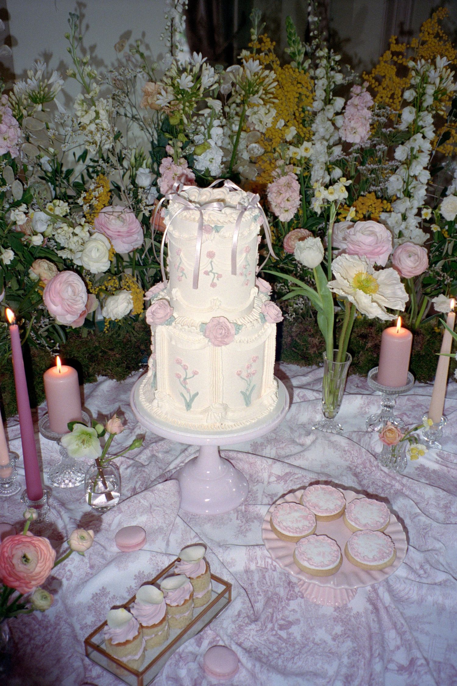 Elegant white wedding cake with floral designs displayed on pink stand with candles and spring flowers on lace tablecloth.