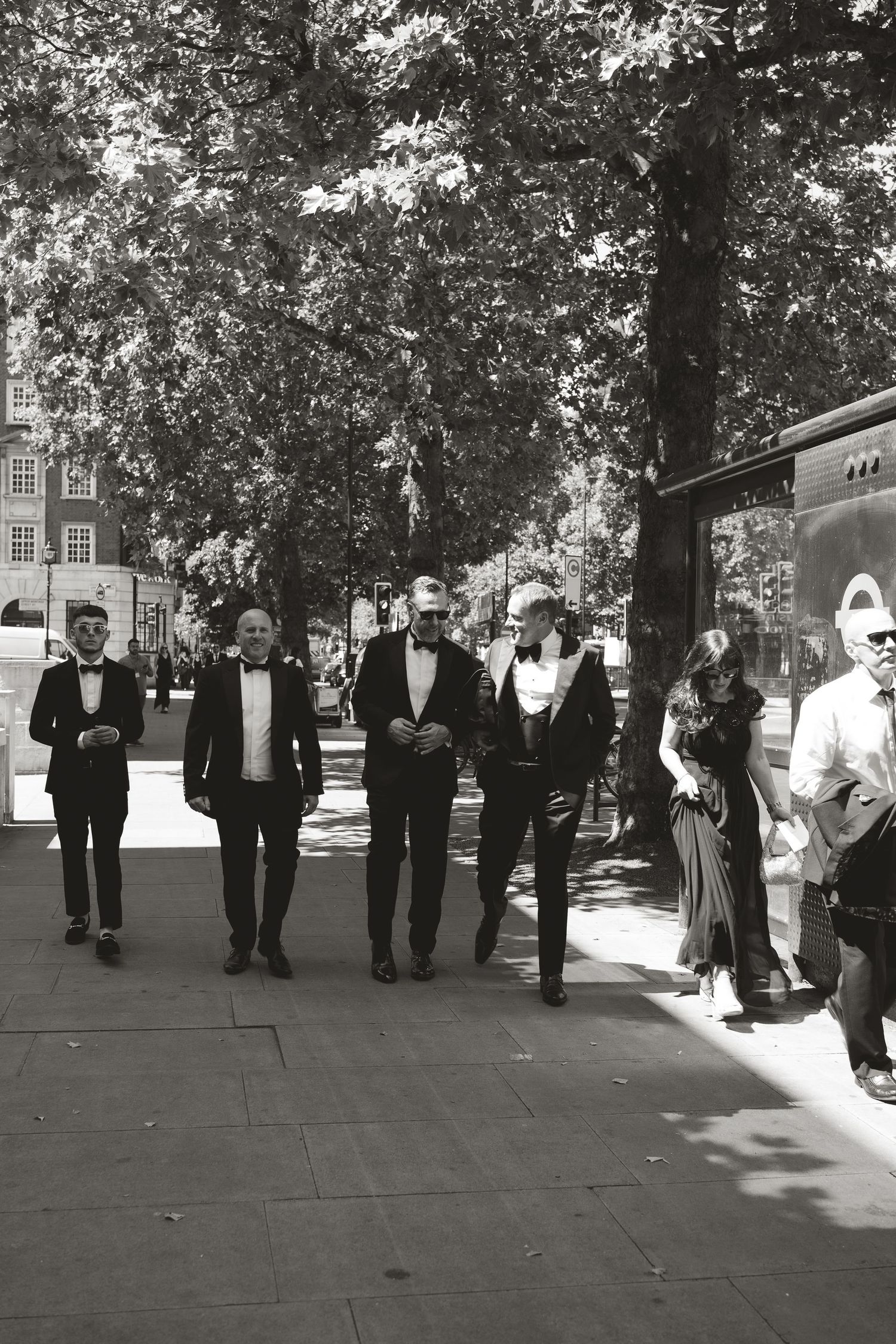 A group of formally dressed people walking together on a tree-lined pathway in black and white.