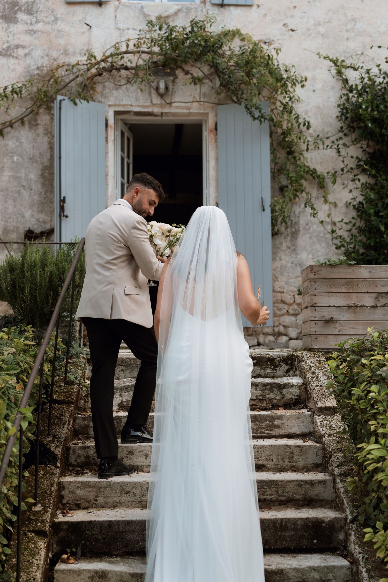 A romantic first look moment between a couple on rustic stone steps with blue shutters and greenery.
