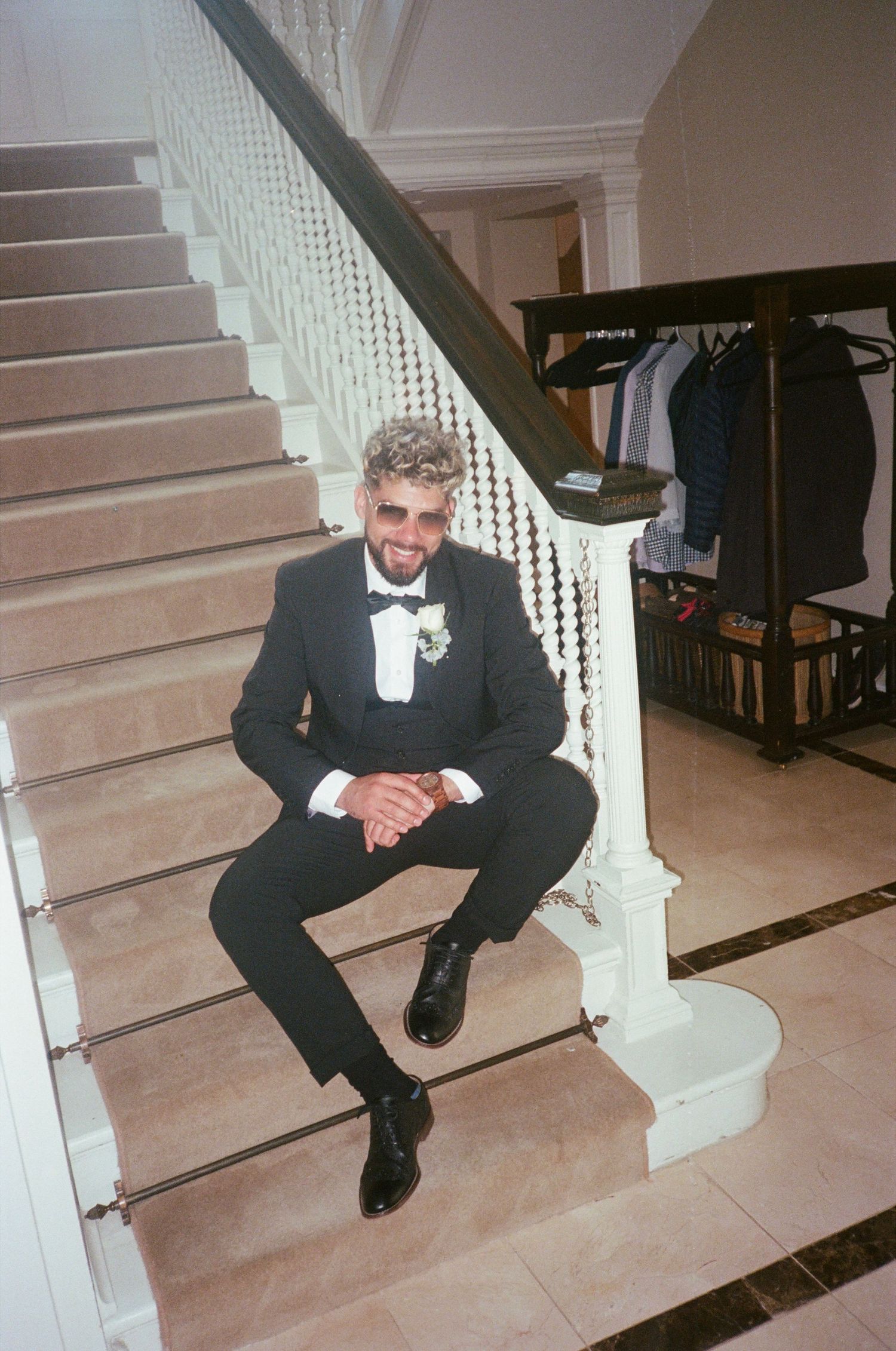 A person in a black tuxedo and bow tie sits on elegant white stairs with a coat rack visible in the background.