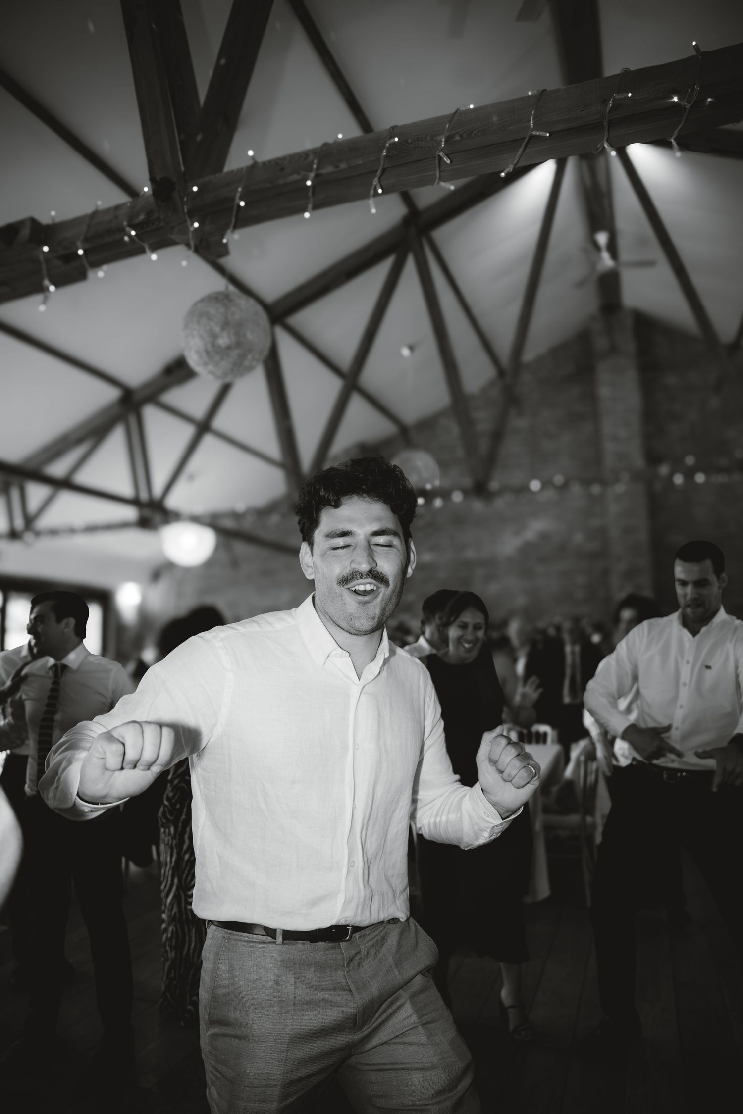 A black and white photo of a wedding guest dancing enthusiastically under string lights.
