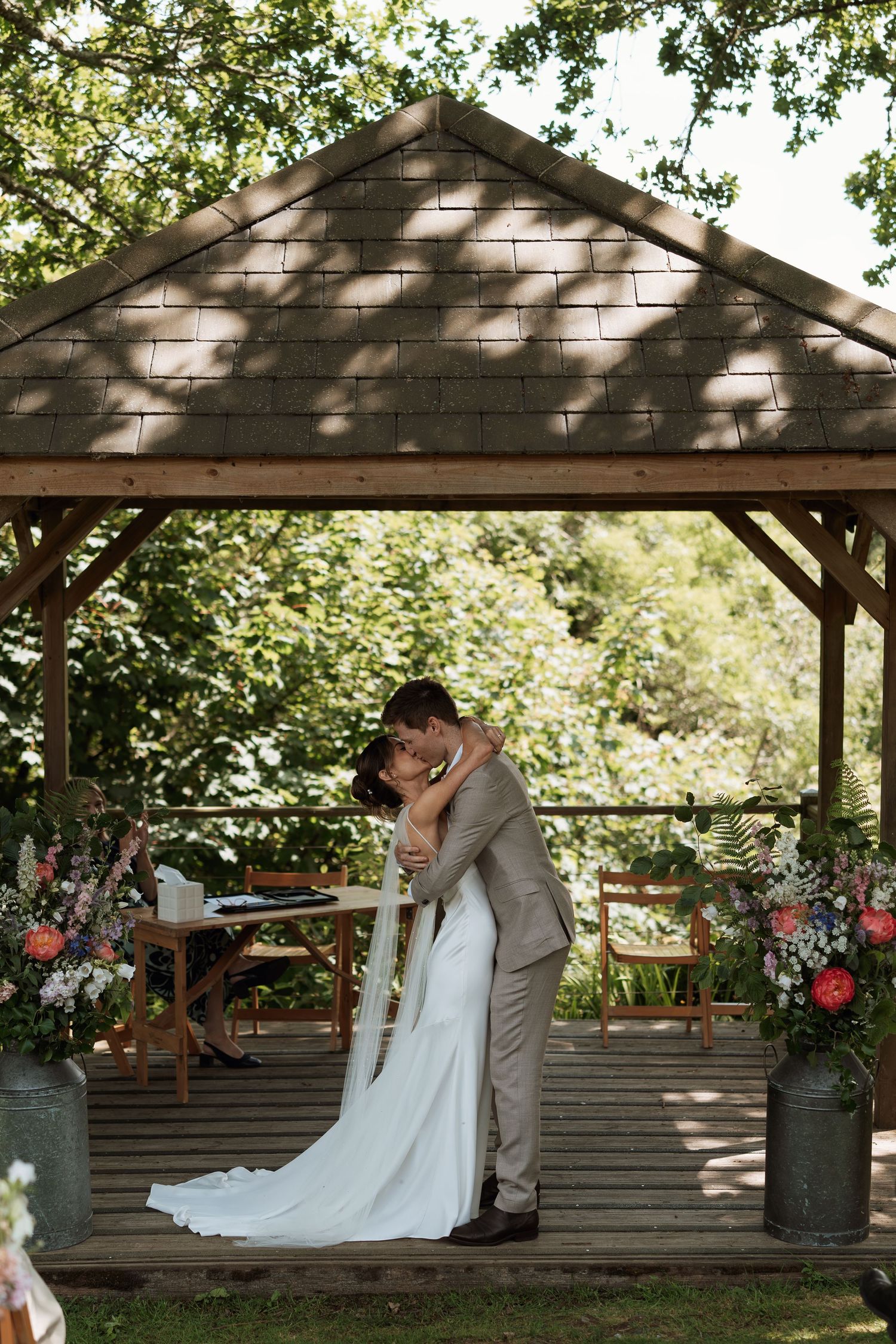A couple sharing a romantic moment under a rustic wooden gazebo decorated with floral arrangements.