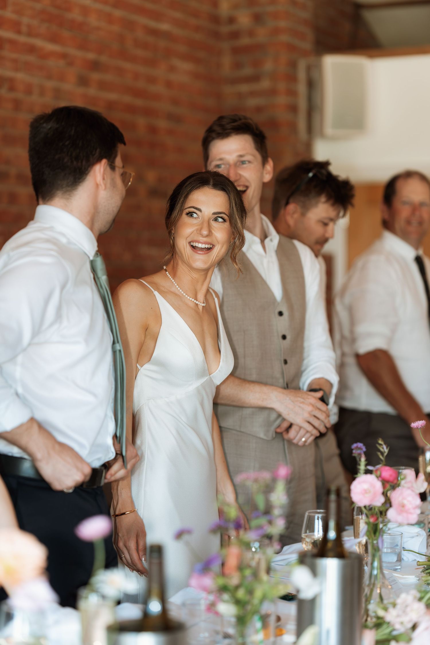Wedding reception scene with guests laughing at decorated tables with pink flowers and brick walls.