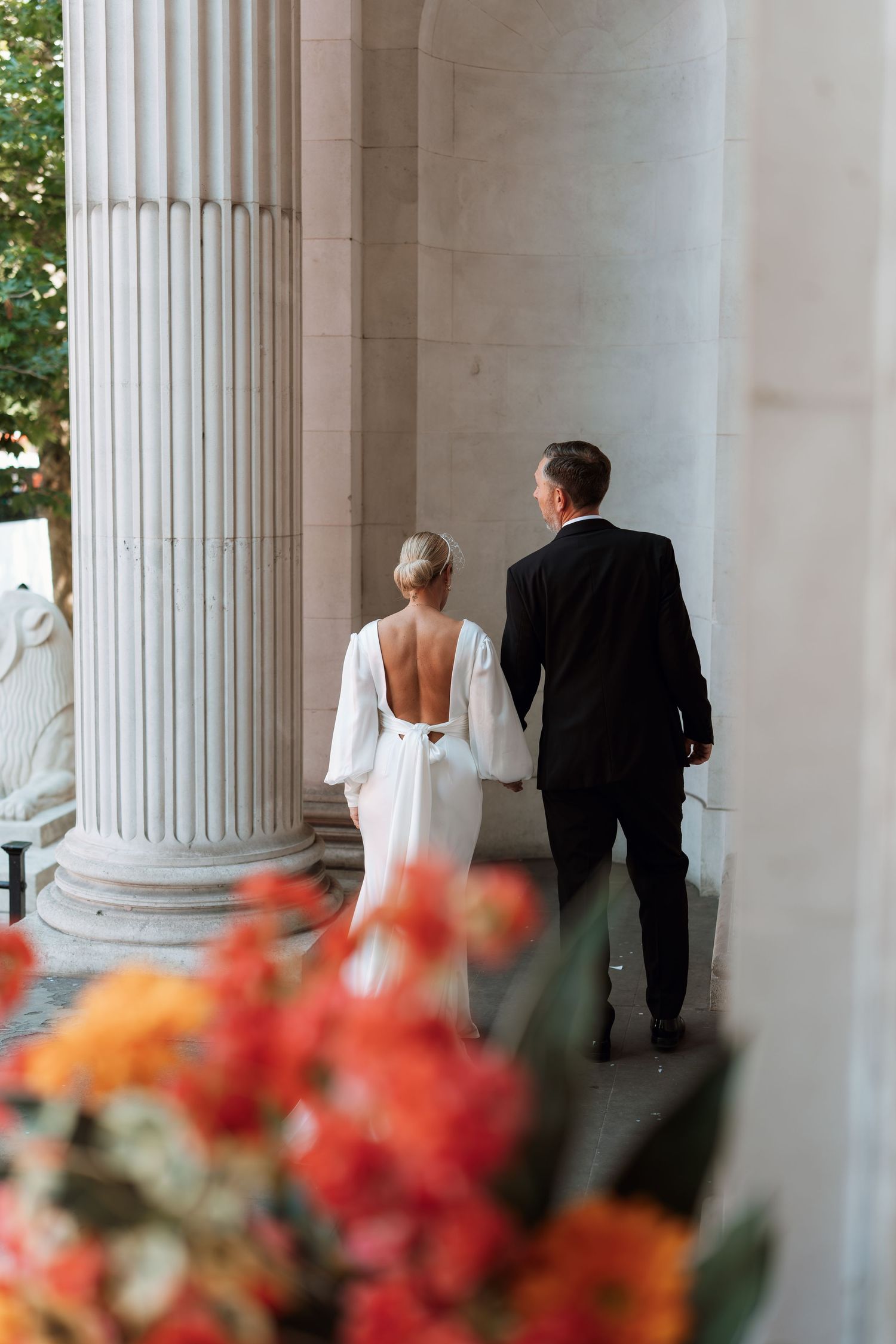 Wedding party walking between elegant white columns with vibrant red and orange flowers in the foreground.