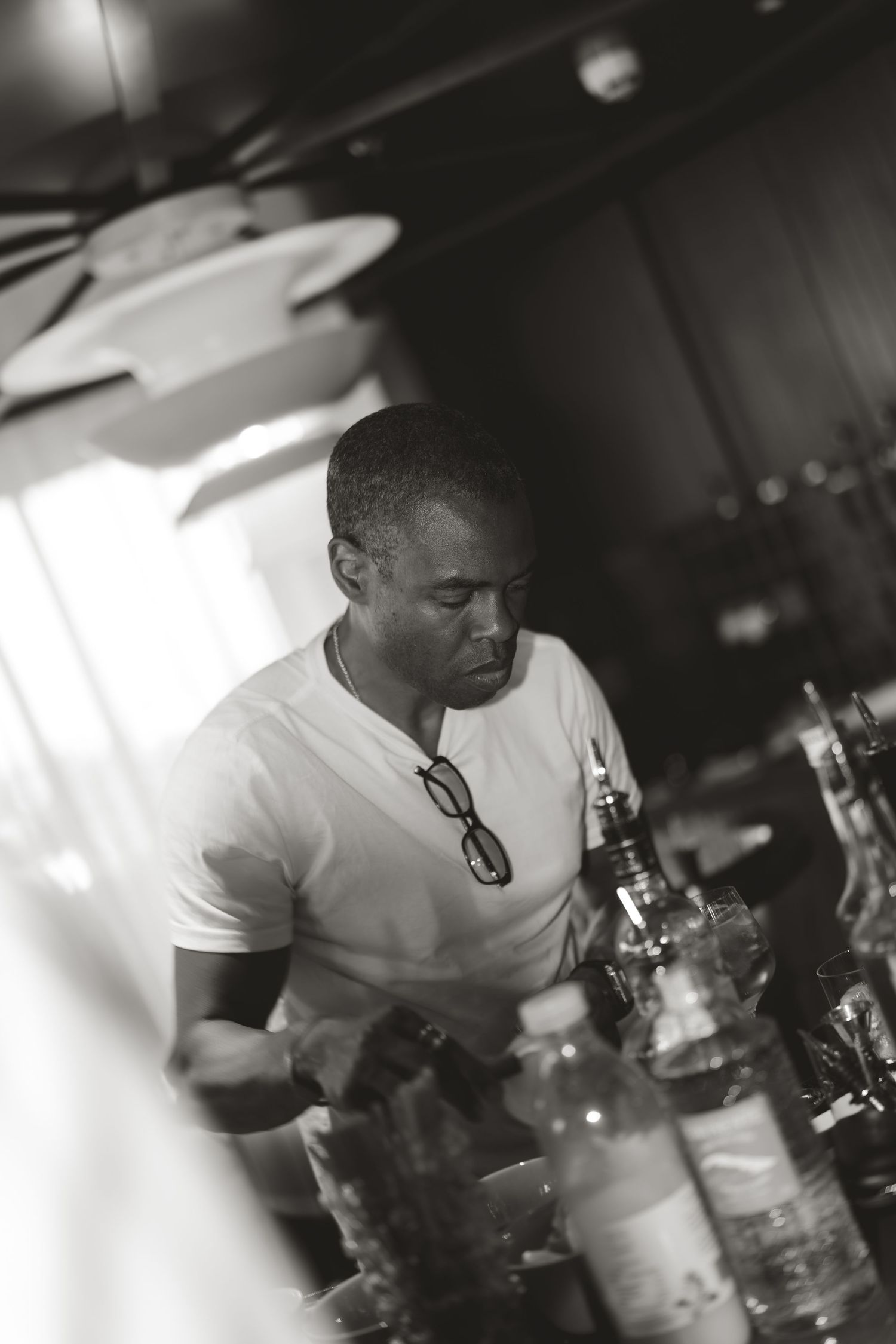Bartender in white t-shirt mixing drinks behind dimly lit bar.