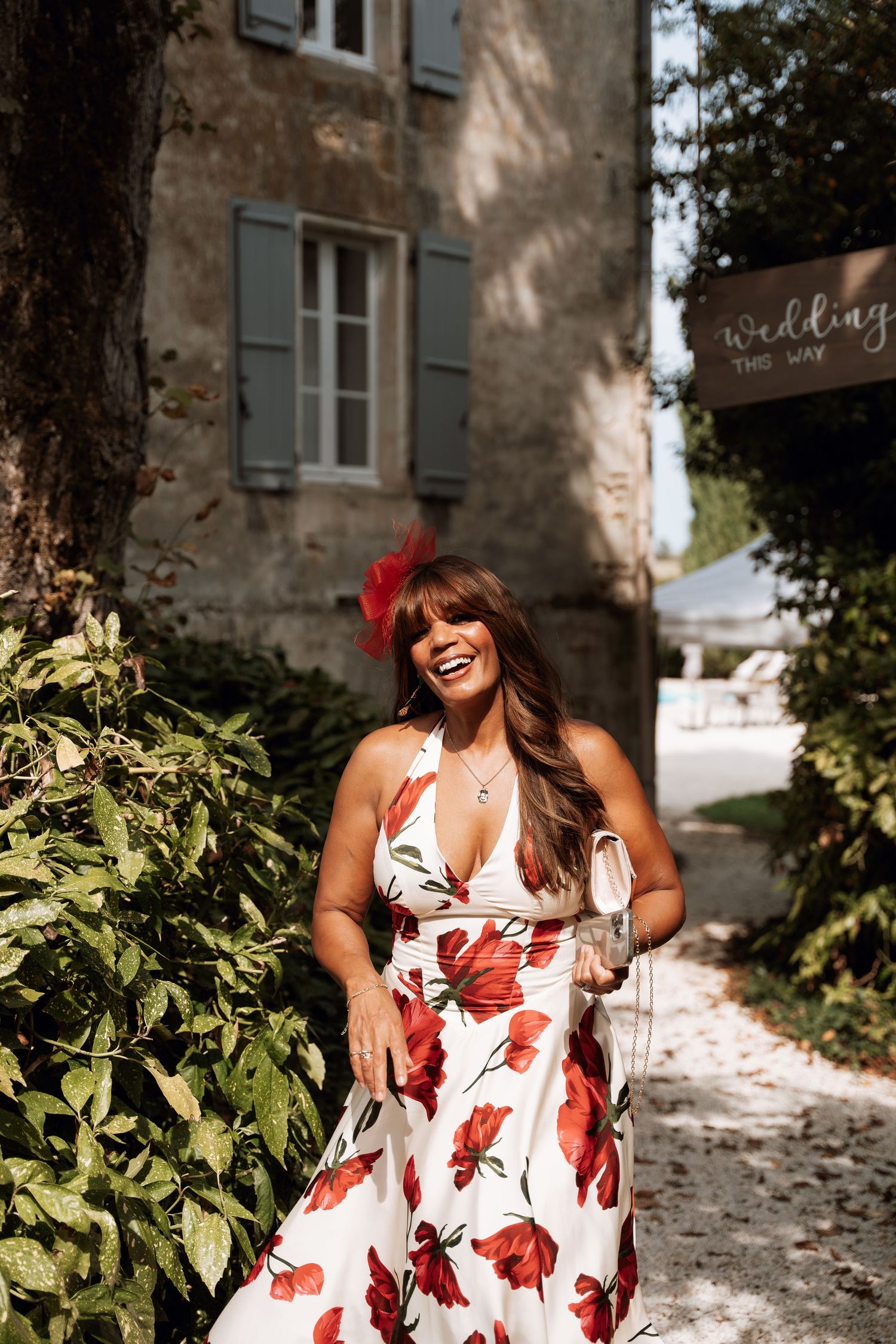 A floral white and red dress is worn against a rustic stone building backdrop with green foliage.