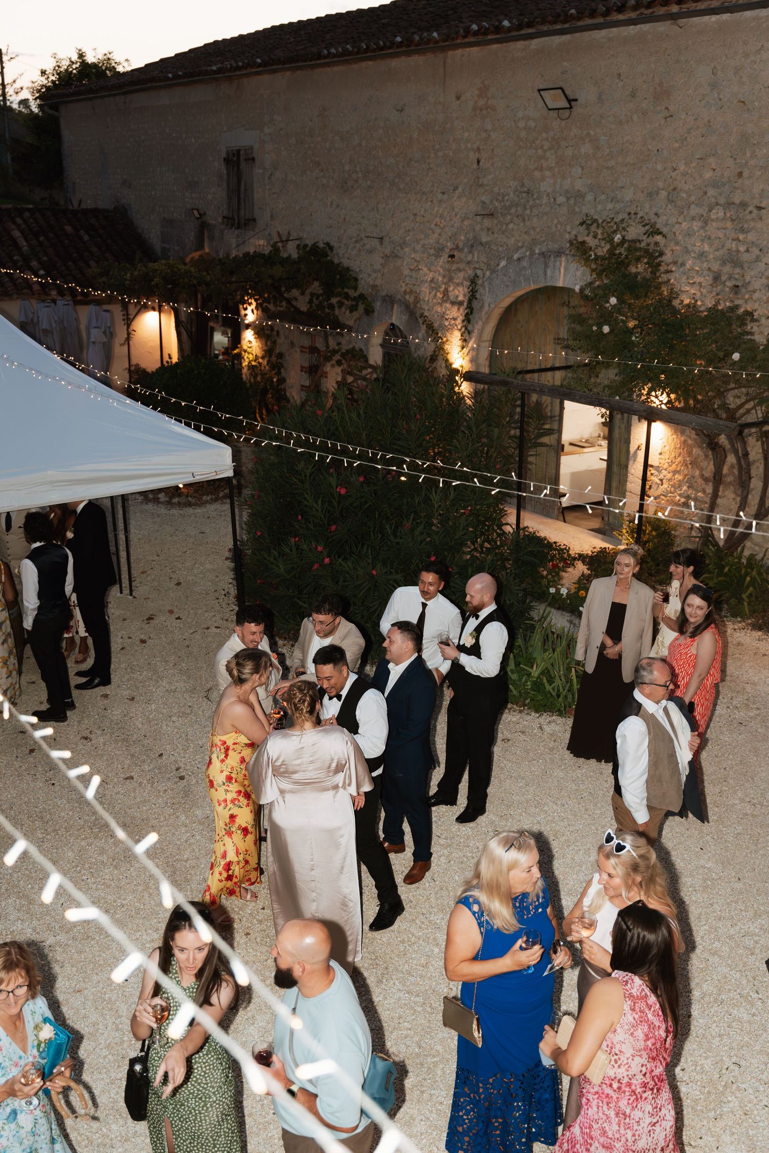 Wedding guests dance under twinkling lights at an outdoor evening reception.