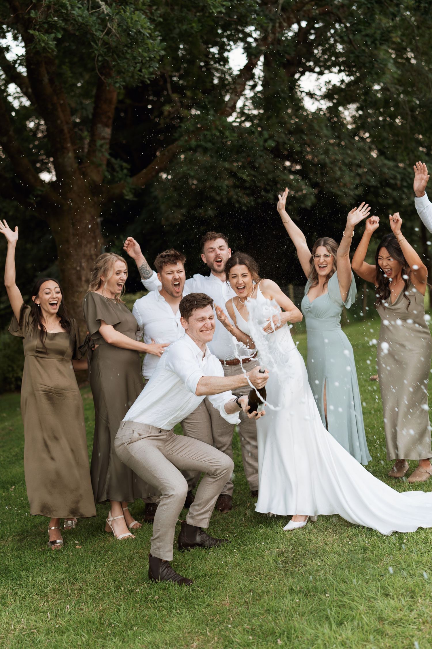 Wedding party jumps for a fun group photo on grassy hill during celebration.