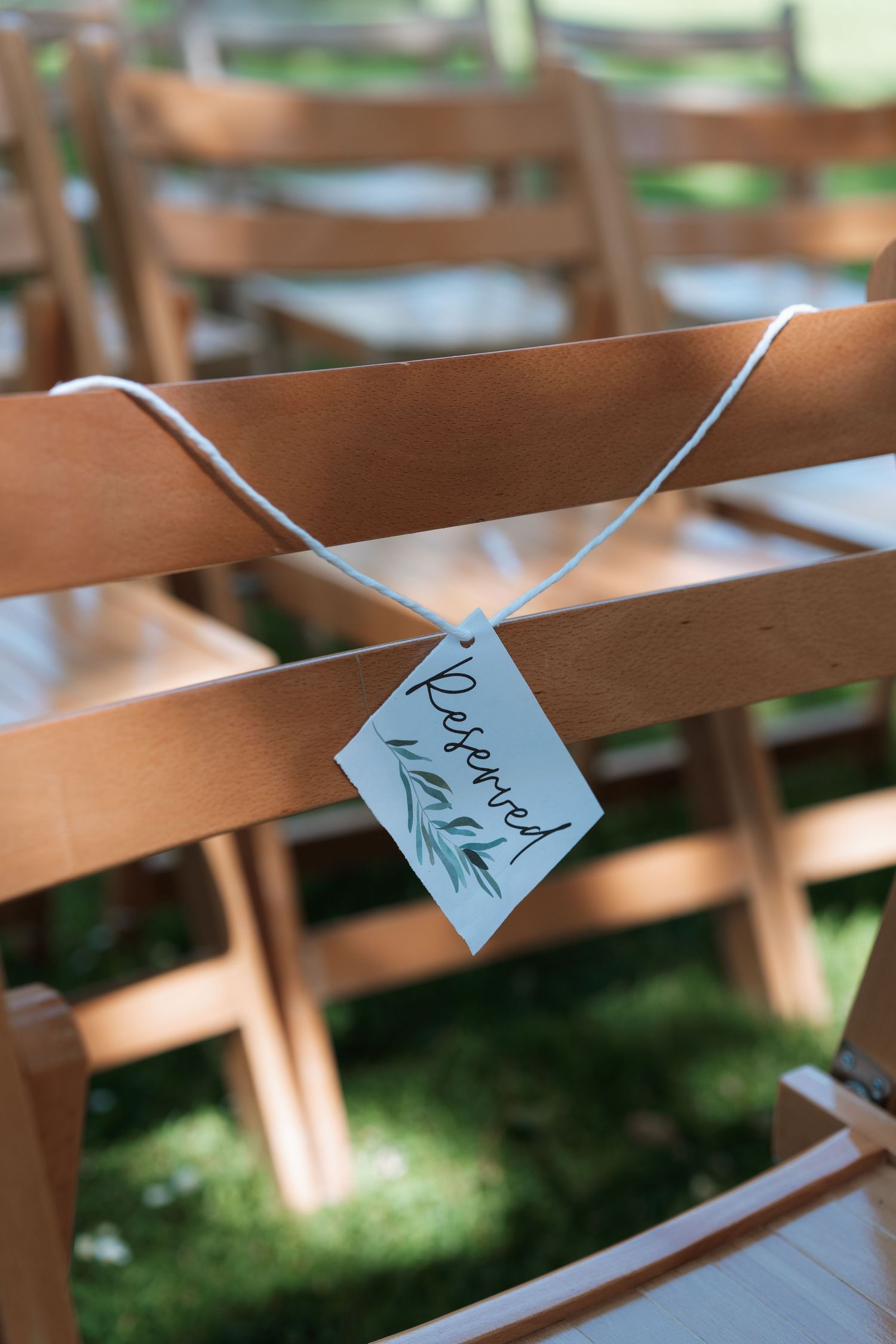 Small paper name cards attached to wooden folding chairs at an outdoor wedding ceremony.