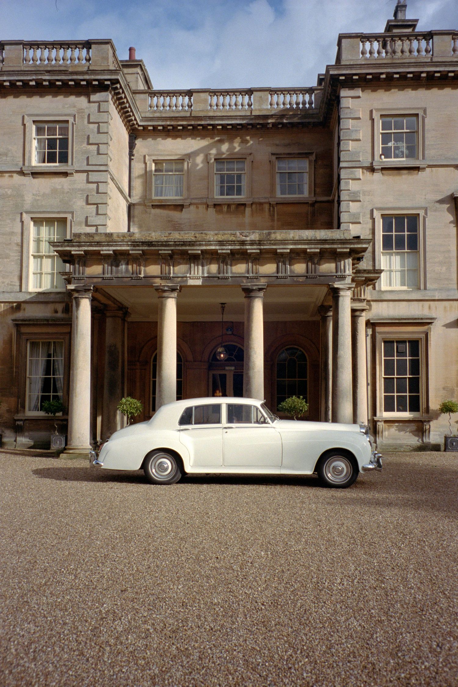 Elegant white vintage Rolls-Royce parked in front of a grand stone mansion with classical columns and balustrades.