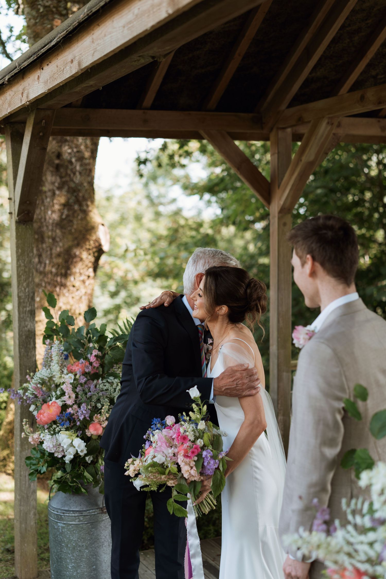 Wedding ceremony taking place under a rustic wooden gazebo decorated with colorful floral arrangements.