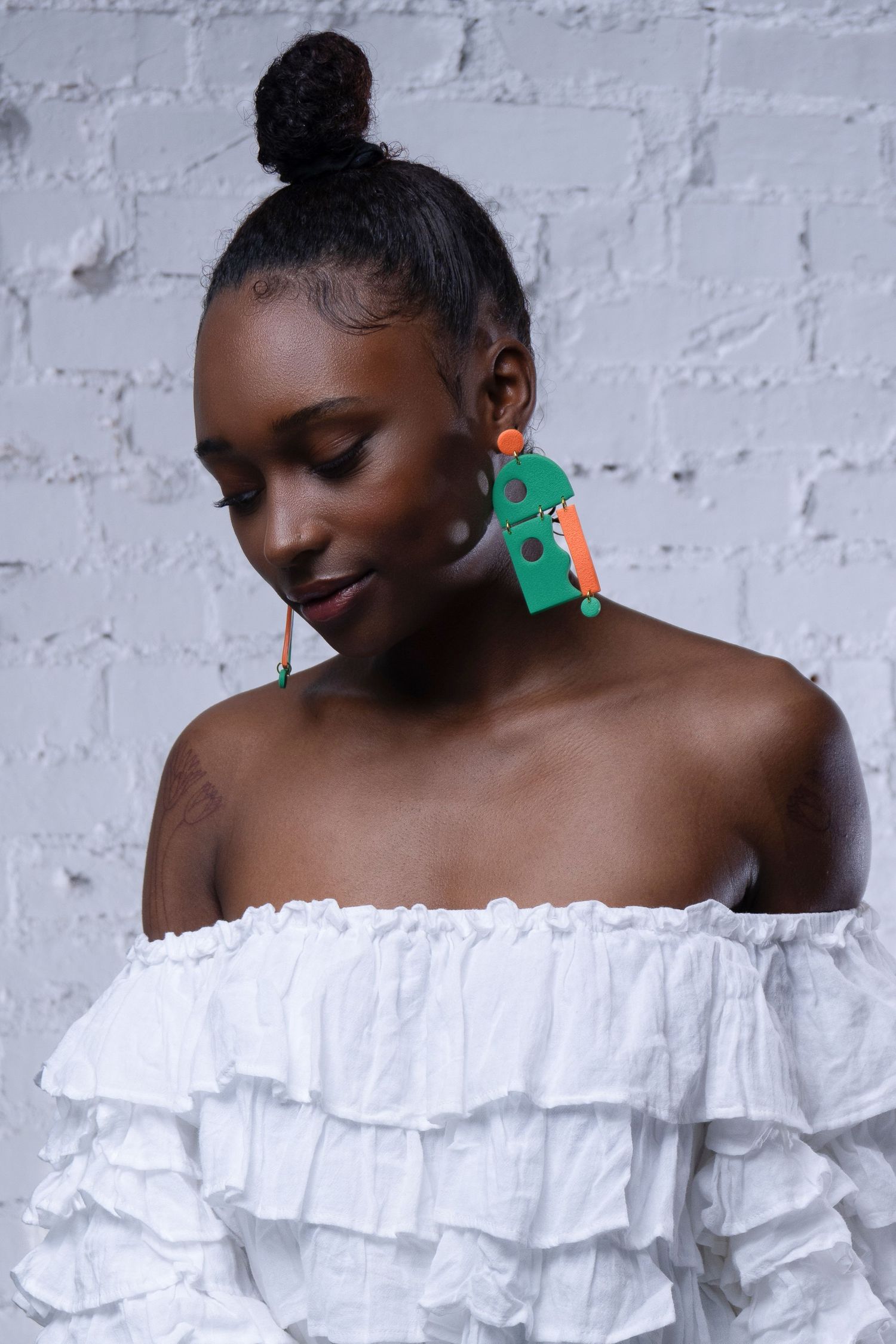 Editorial portrait of model in white ruffled blouse with bold geometric statement earrings against textured brick backdrop.