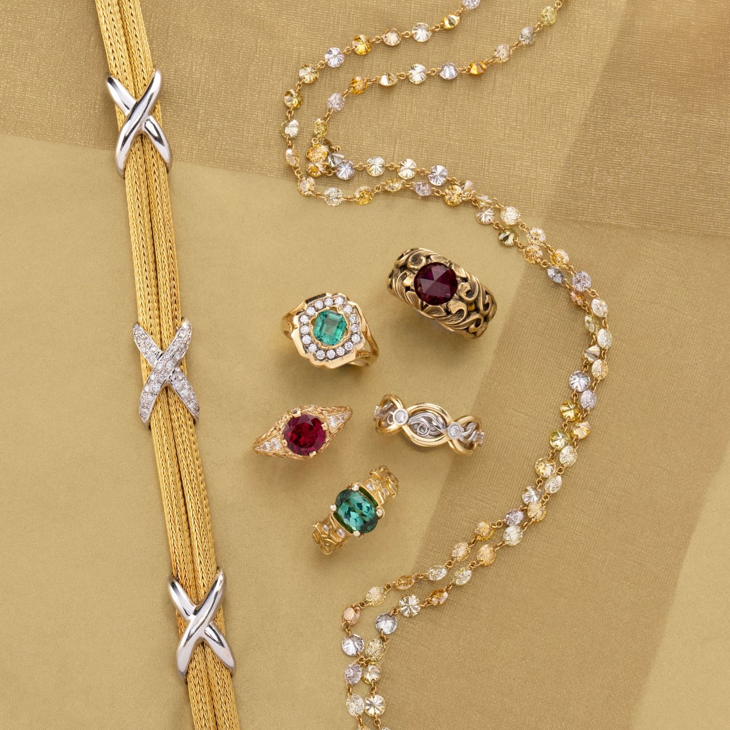 Gold rings with emerald and ruby stones styled with layered chains on a warm, elegant studio background.