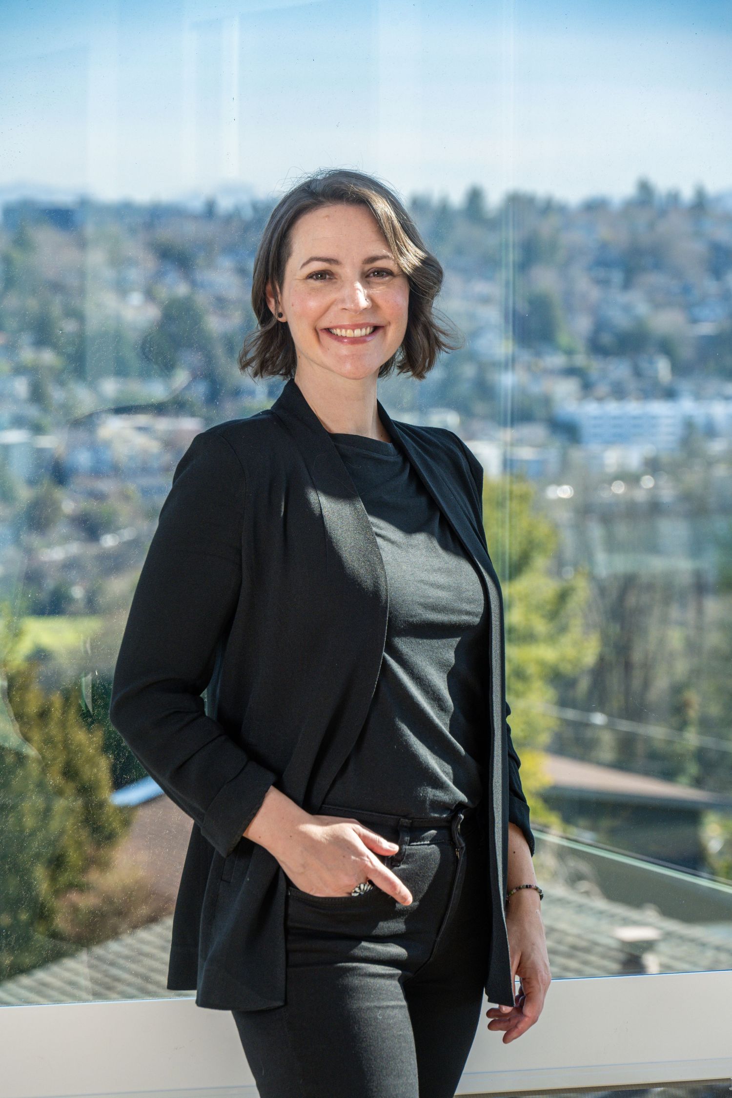 Professional business portrait of woman standing by window with city view, photographed in natural light for executive branding.