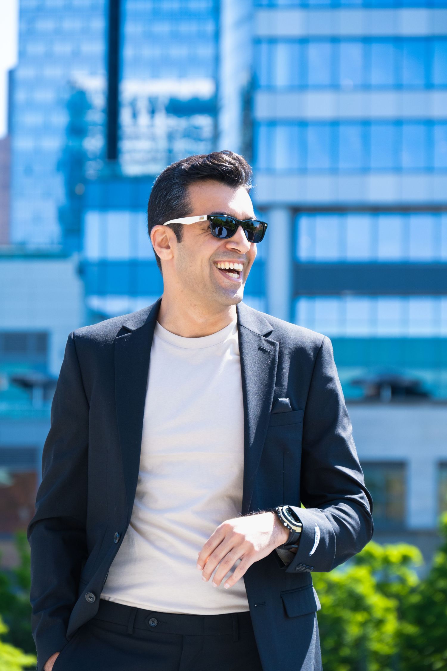 Man in navy blazer and beige t-shirt smiling while wearing sunglasses, standing outdoors with modern glass buildings in background.