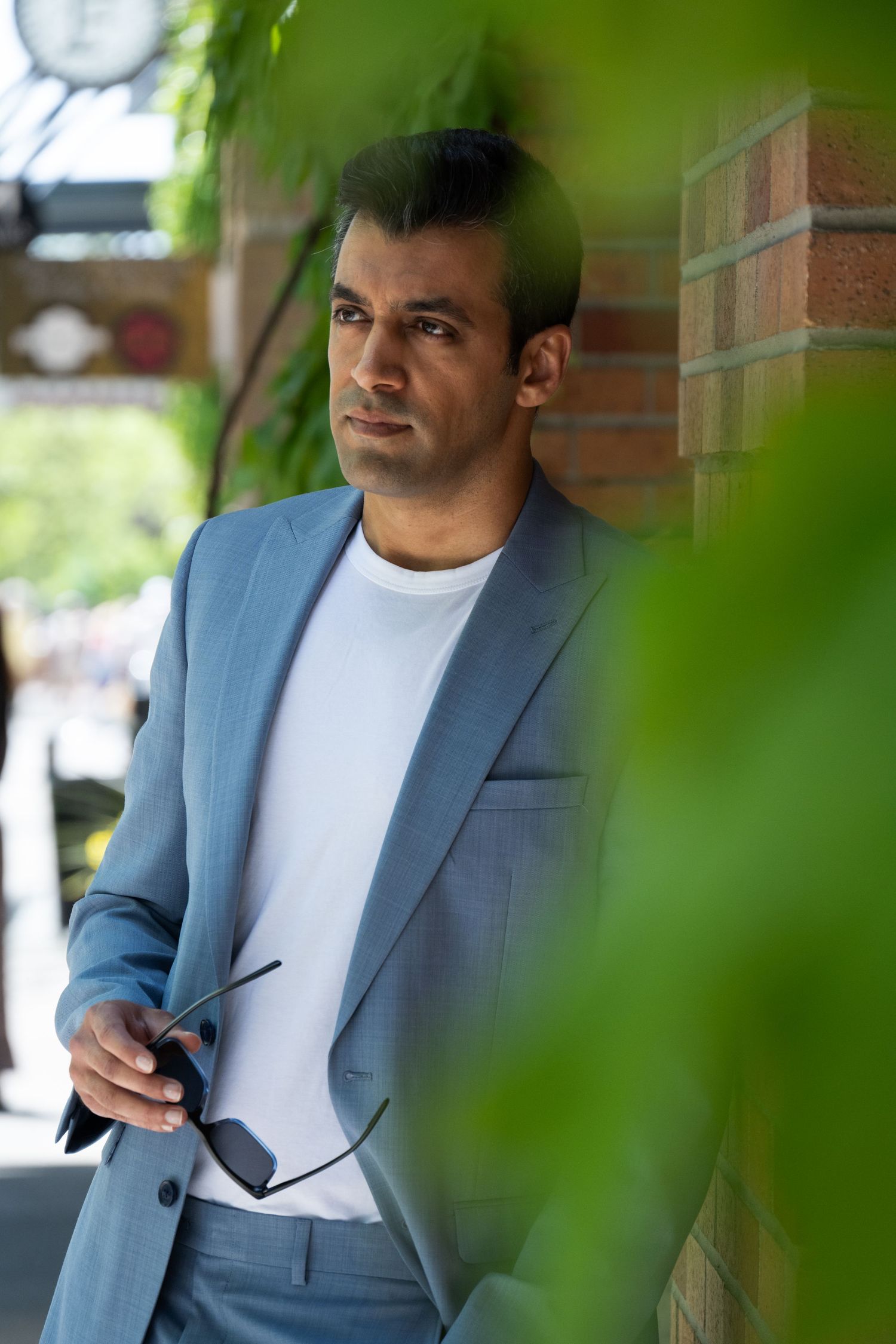 Man in light blue tailored suit and white t-shirt leaning against brick wall outdoors, holding sunglasses, framed by soft green leaves.