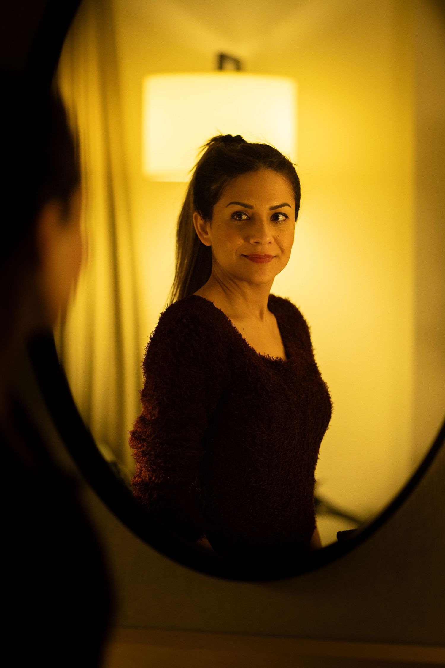 Lifestyle portrait of woman reflected in mirror with warm ambient lighting, creating an intimate and cinematic mood.