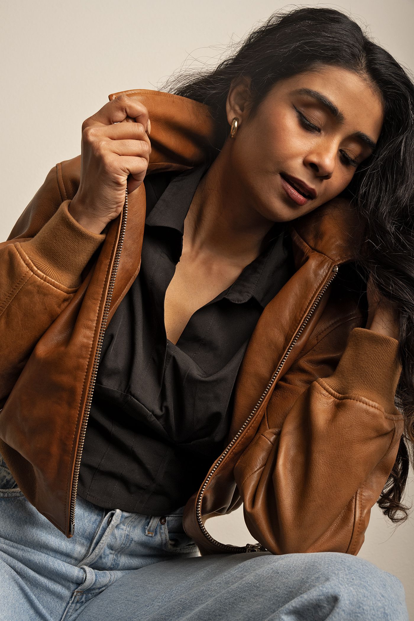 Editorial fashion portrait of woman in brown leather jacket and black blouse, soft studio lighting.