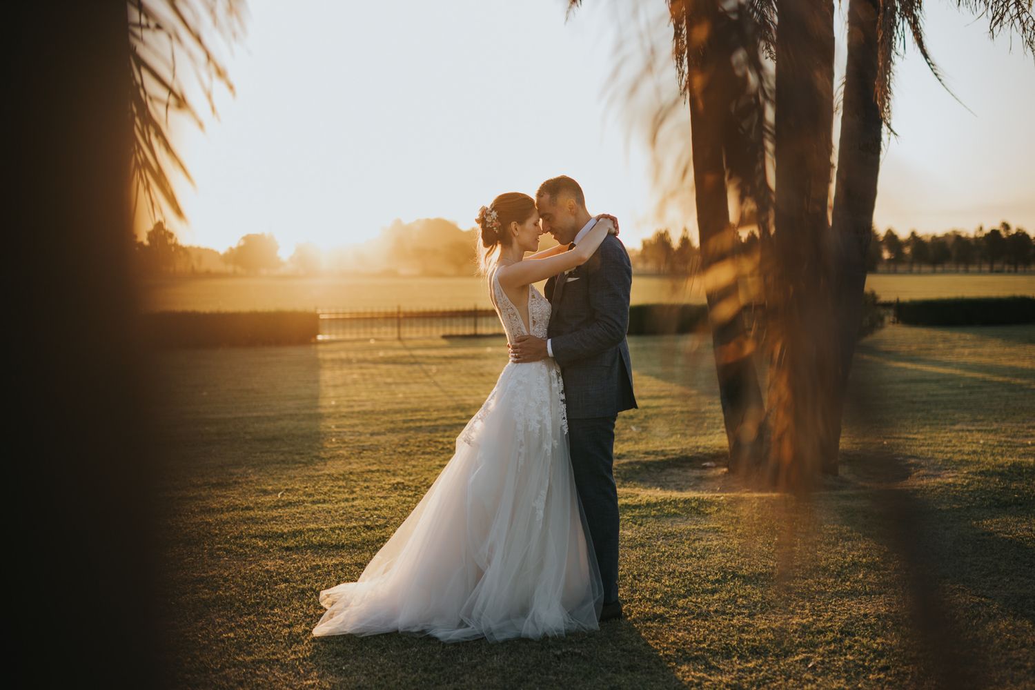 Bride and groom embrace at golden hour sunset on a grassy field framed by palm trees, the bride in a tulle wedding gown.