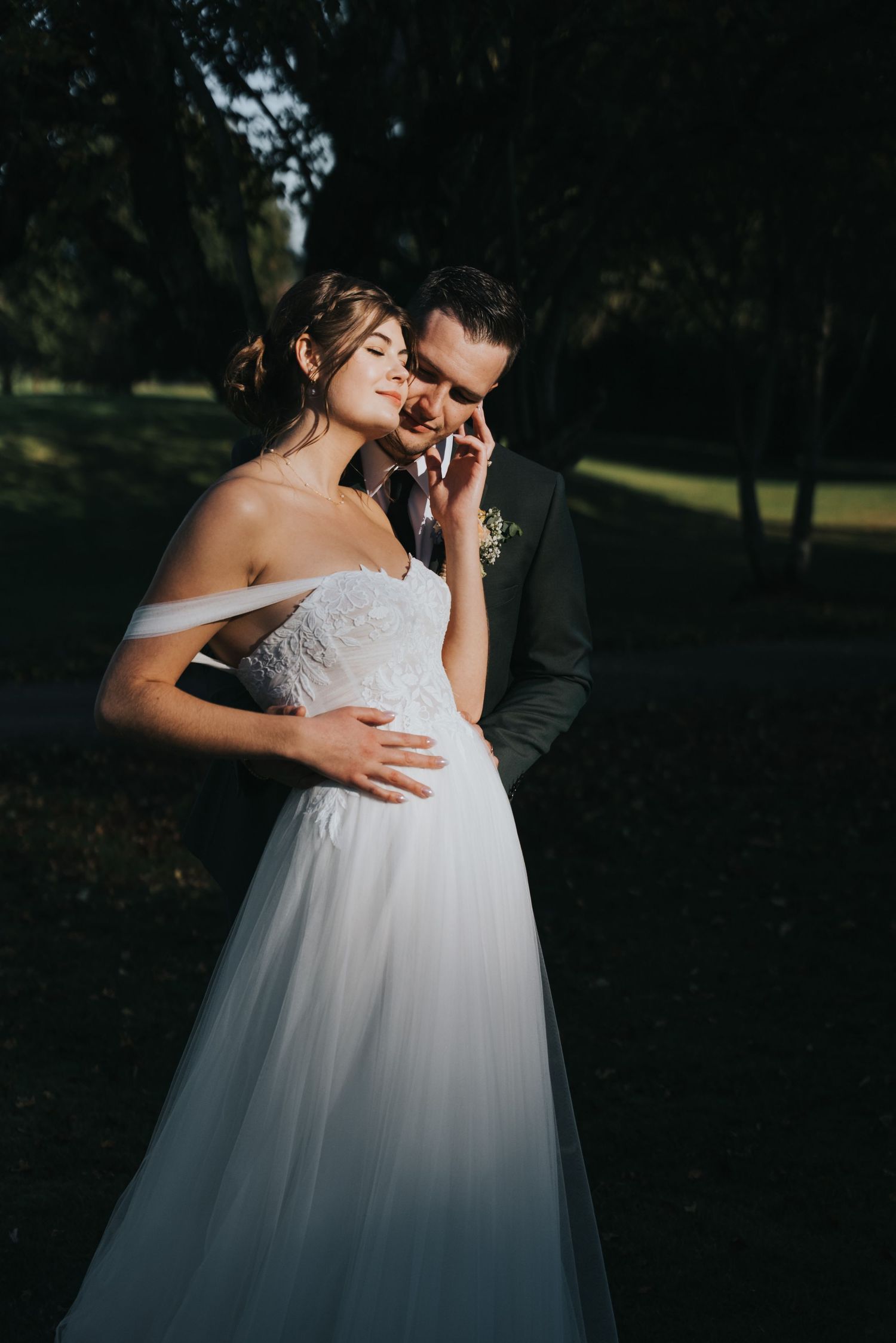 Bride in an off-shoulder white tulle wedding gown embraces her groom in a dark suit with a boutonniere in a sunlit, tree-lined garden.