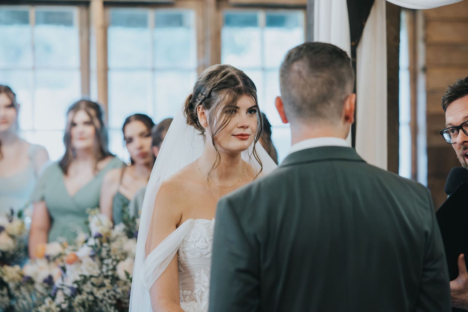Bride in a white lace gown and veil faces her groom in a gray suit during a wedding ceremony as bridesmaids look on.