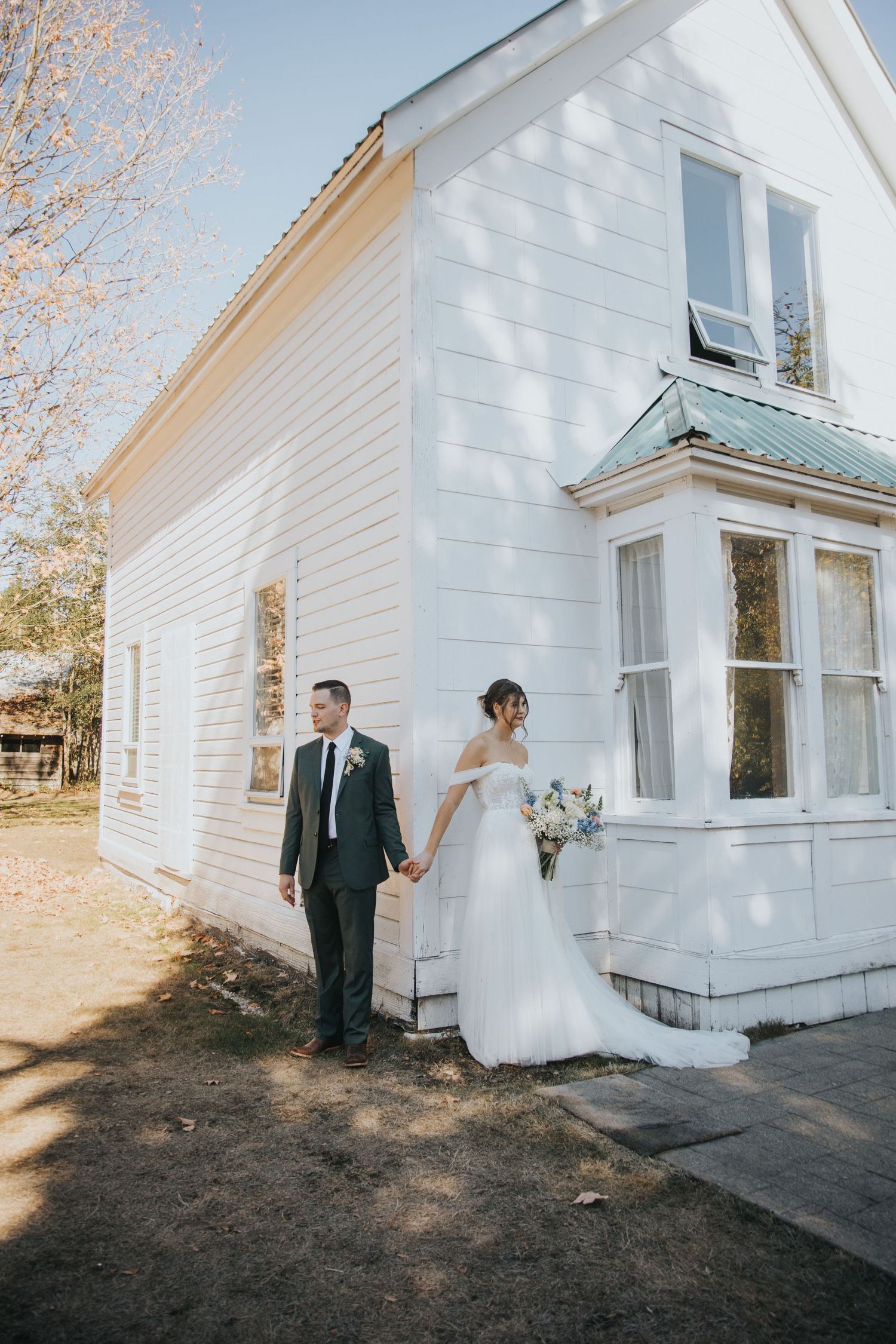 Bride in an off-shoulder gown and groom in a dark suit hold hands outside a white wooden building on their wedding day.