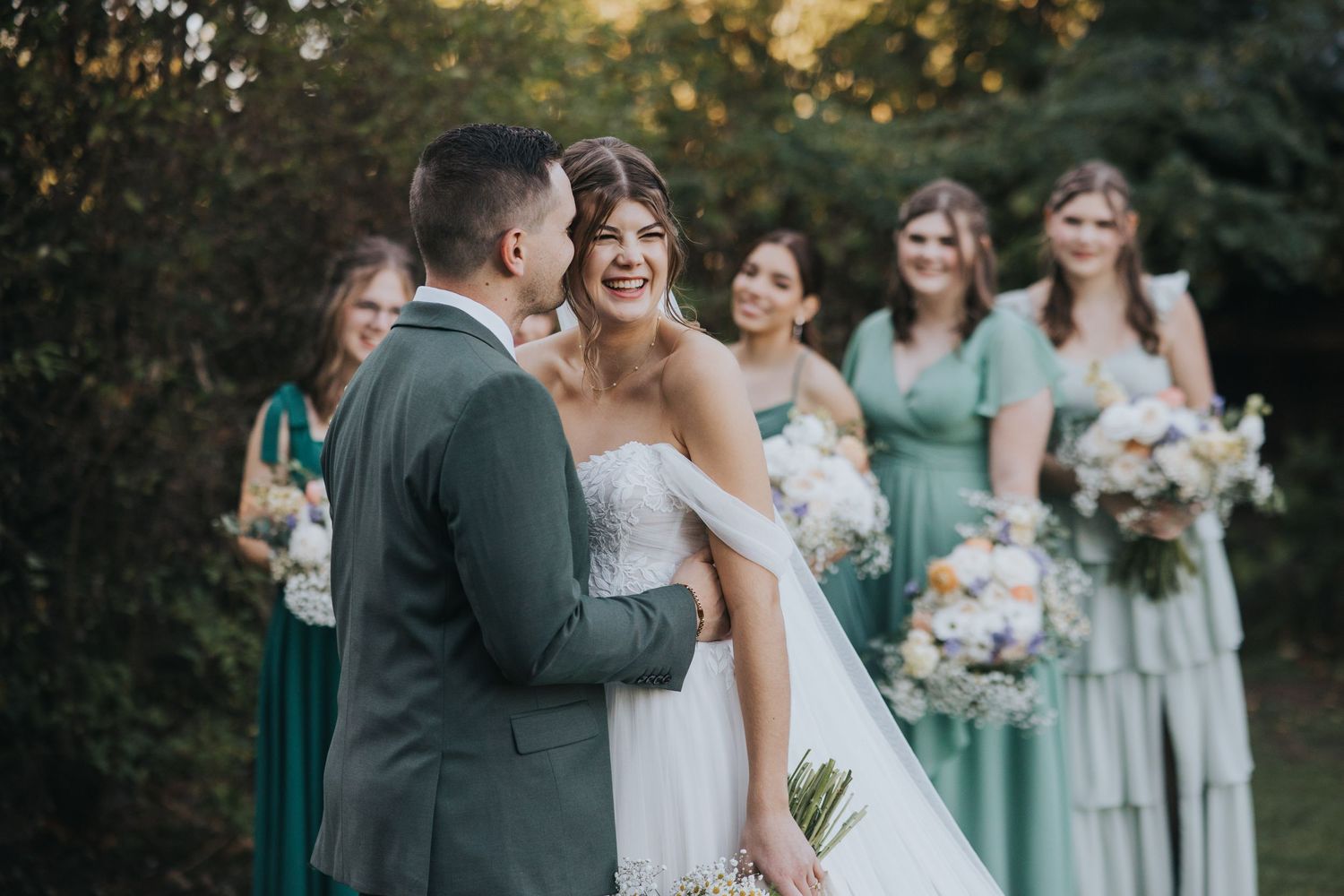 Bride laughing in the groom's arms with bridesmaids in sage dresses behind them