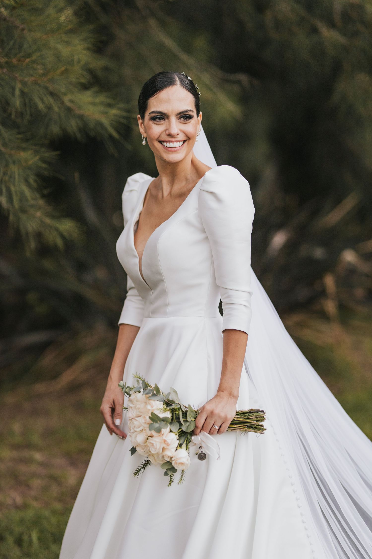 Smiling bride in an elegant long-sleeve satin wedding gown and veil holding a bouquet of cream roses outdoors.