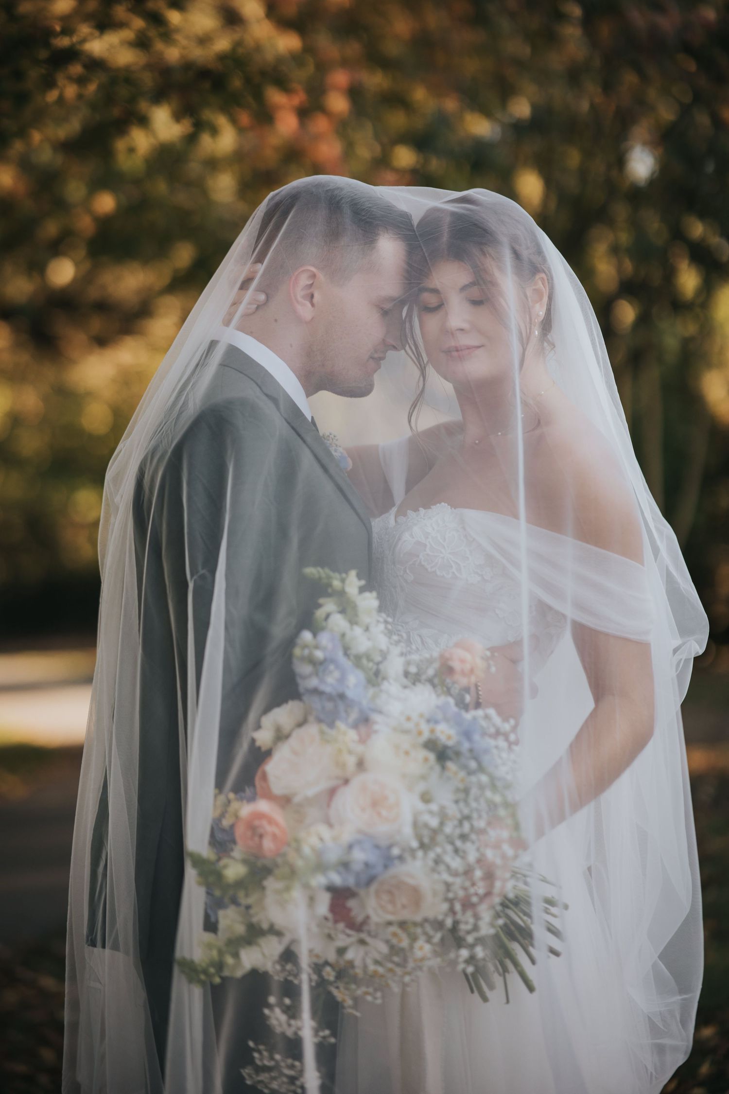Bride and groom with foreheads together under her veil in golden autumn light