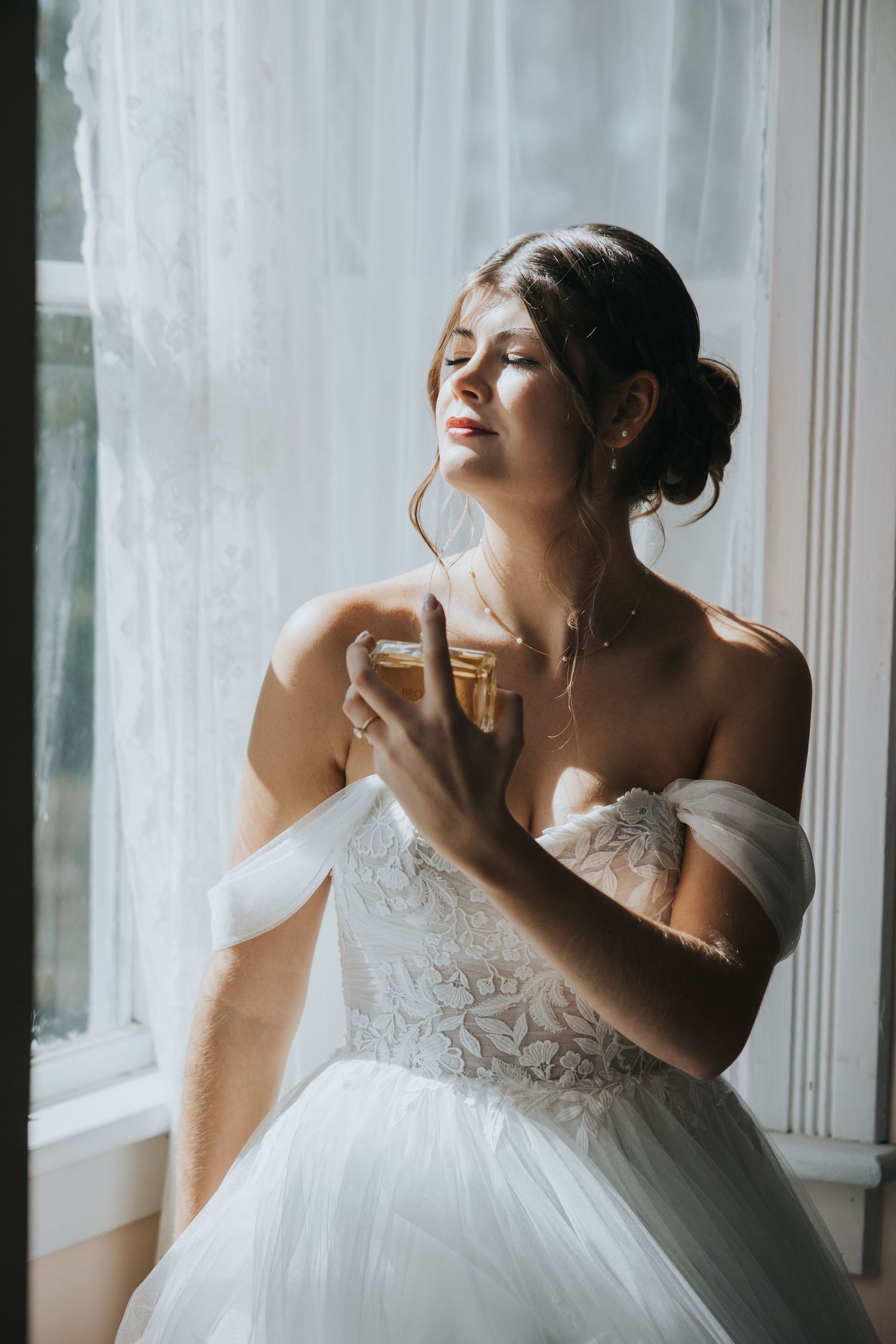 Bride in an off-the-shoulder lace wedding gown with closed eyes holds perfume by a sunlit window with sheer curtains.