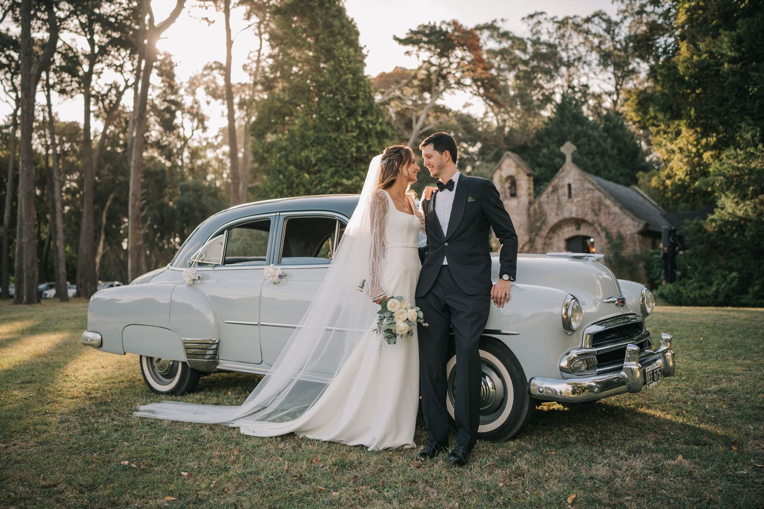Bride in a flowing white gown and groom in a black tuxedo embrace beside a vintage car on a sunny lawn near a stone chapel.