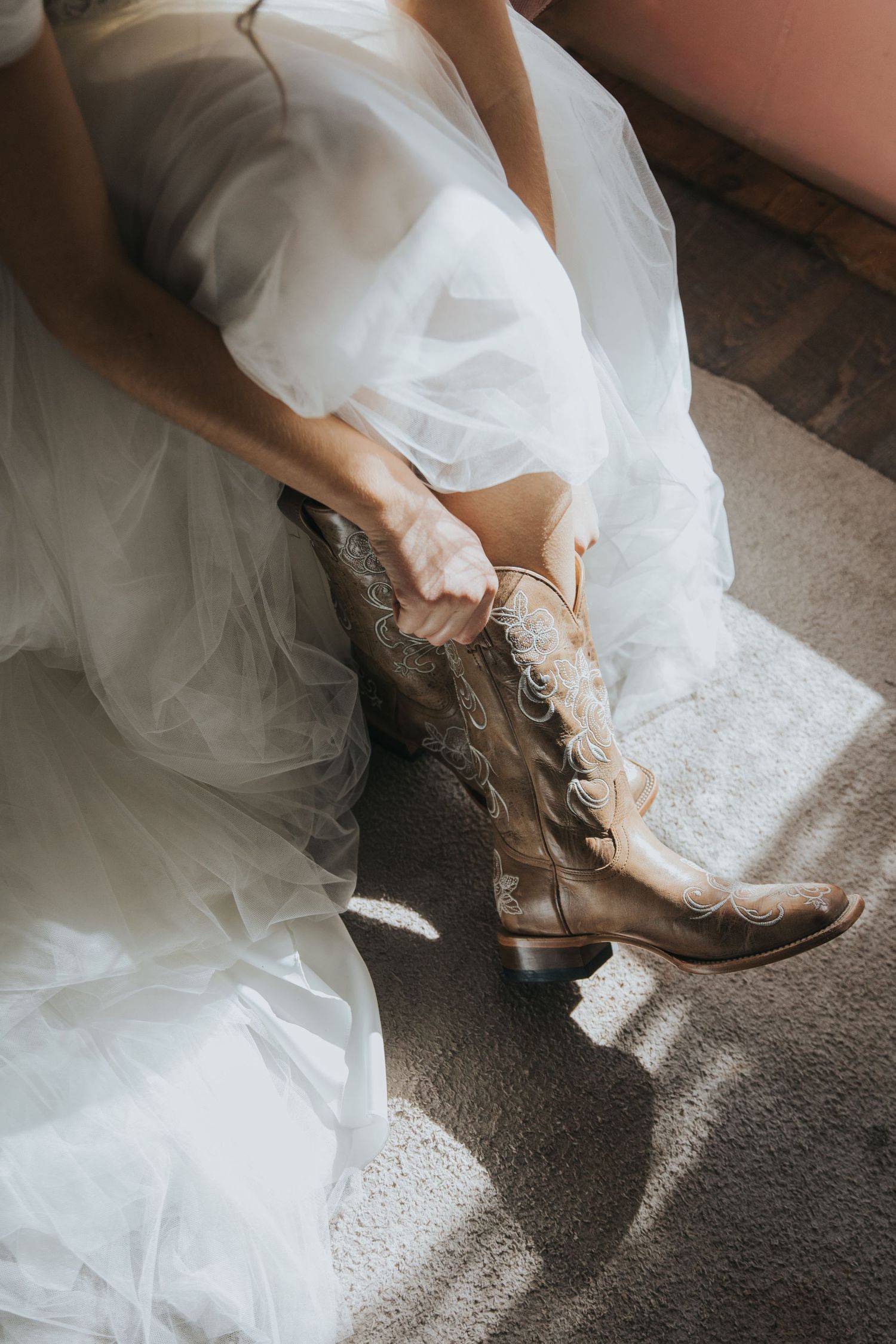 Bride in a white tulle wedding dress pulling on tan embroidered cowboy boots in a sunlit room.