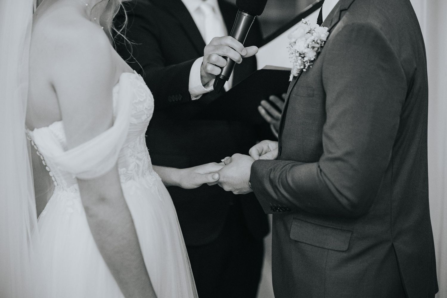 Black and white photo of a bride and groom holding hands as an officiant with a microphone leads their wedding ceremony.