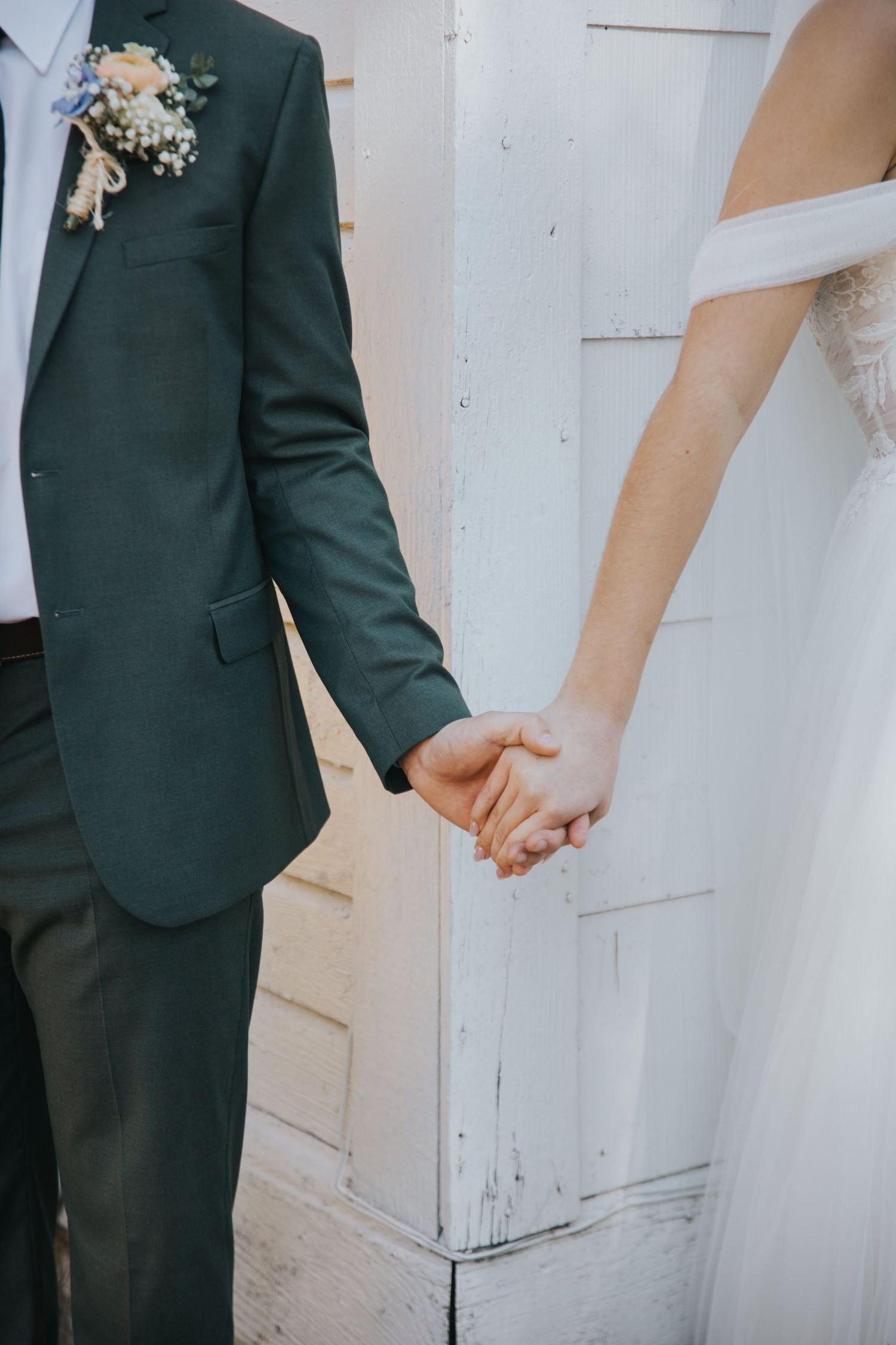 Bride and groom holding hands beside a white wooden wall, the groom in a green suit with a floral boutonniere and the bride in a tulle gown.