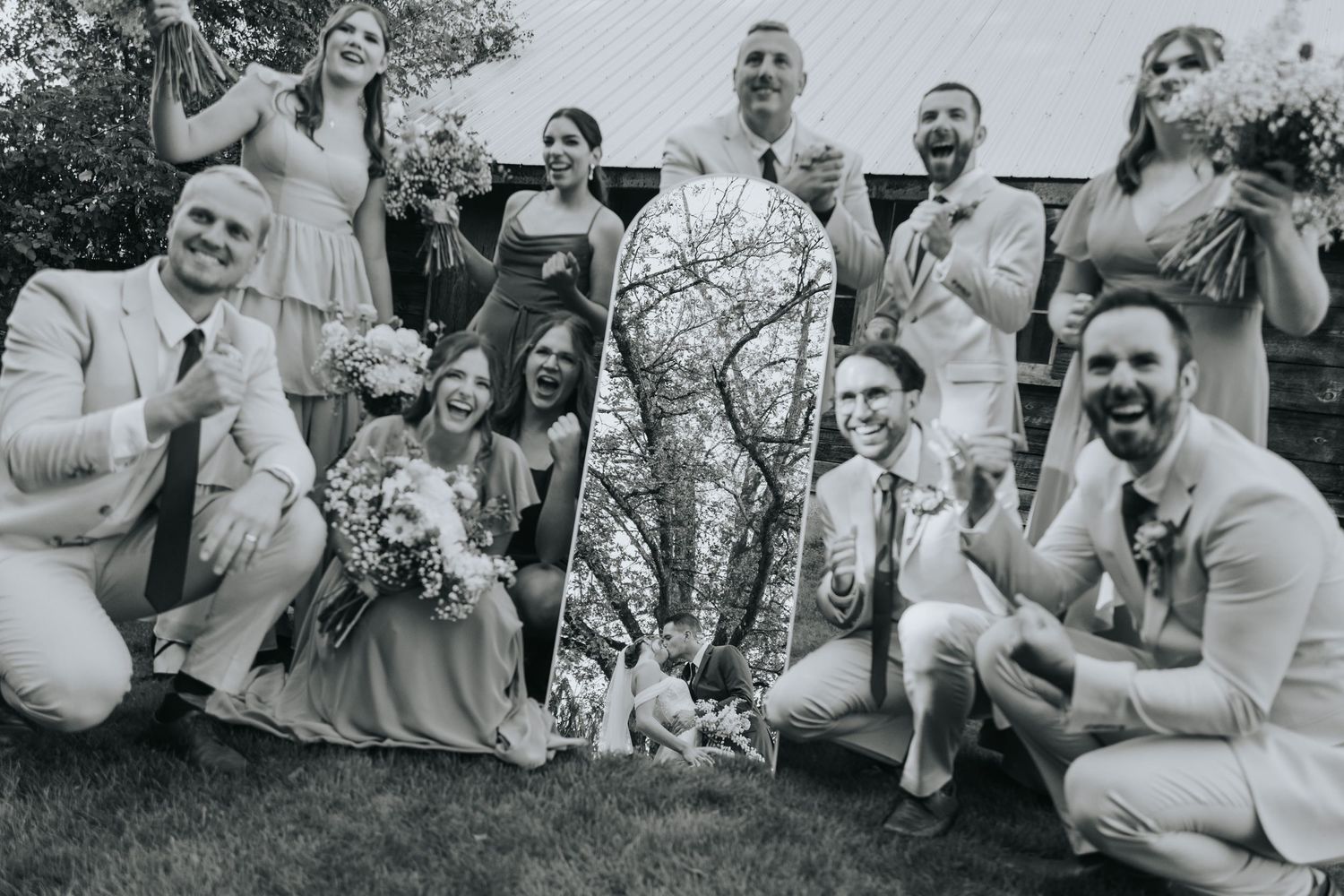 Black and white photo of the bridal party cheering around a mirror reflecting the couple kissing