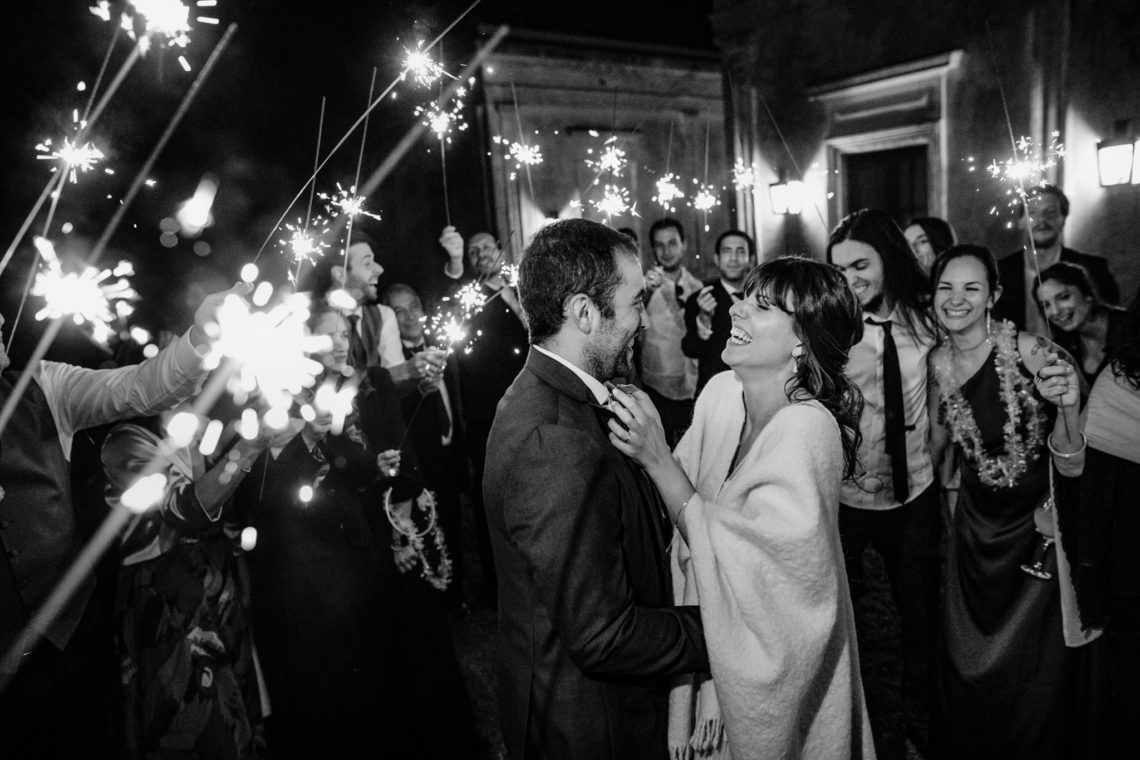 Bride and groom share a joyful moment as wedding guests hold glowing sparklers in a black and white photo.
