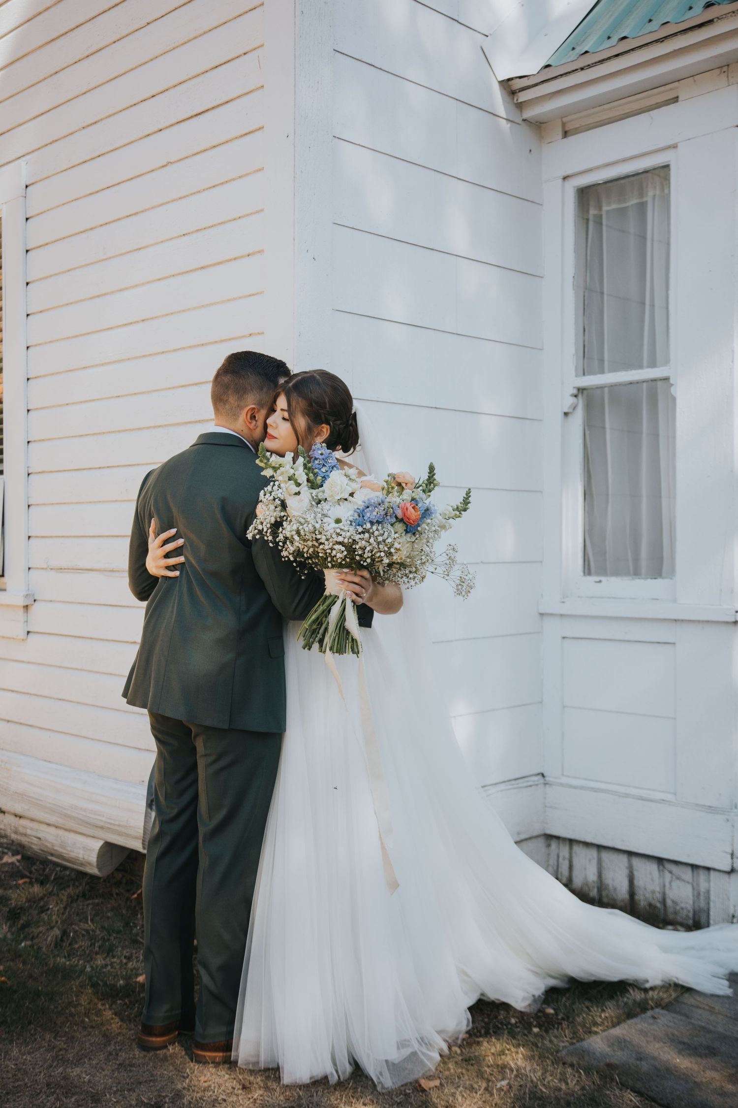 Bride in a flowing tulle gown holding a blue and white bouquet embraces a groom in a green suit beside a white wooden building.