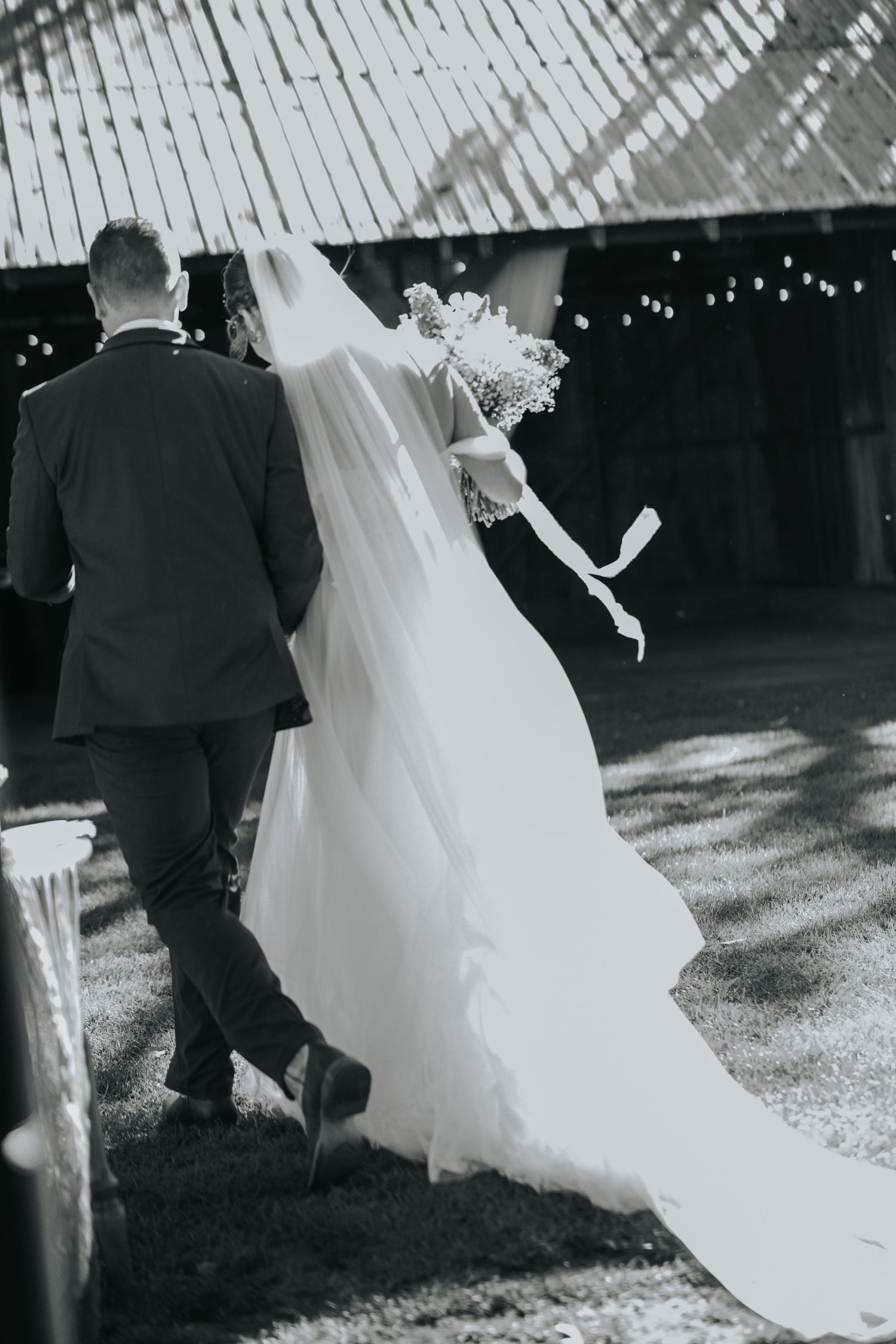 Black and white photo of the couple walking toward the barn, her long veil trailing behind