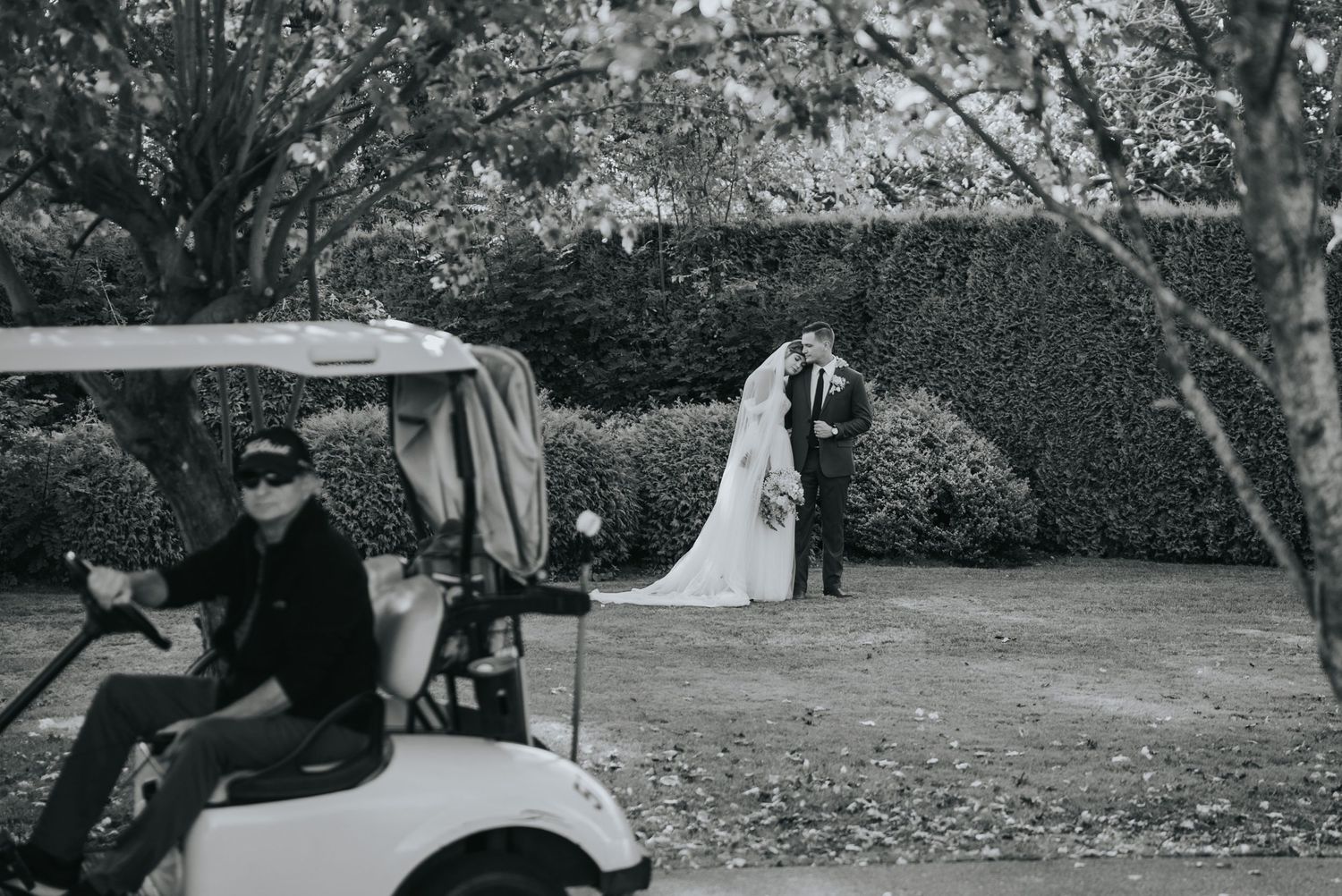 Black and white photo of the couple embracing by the hedges as a golf cart passes in the foreground