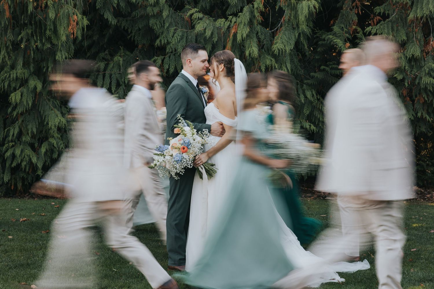 Couple standing still while the bridal party walks around them in motion blur under cedar trees