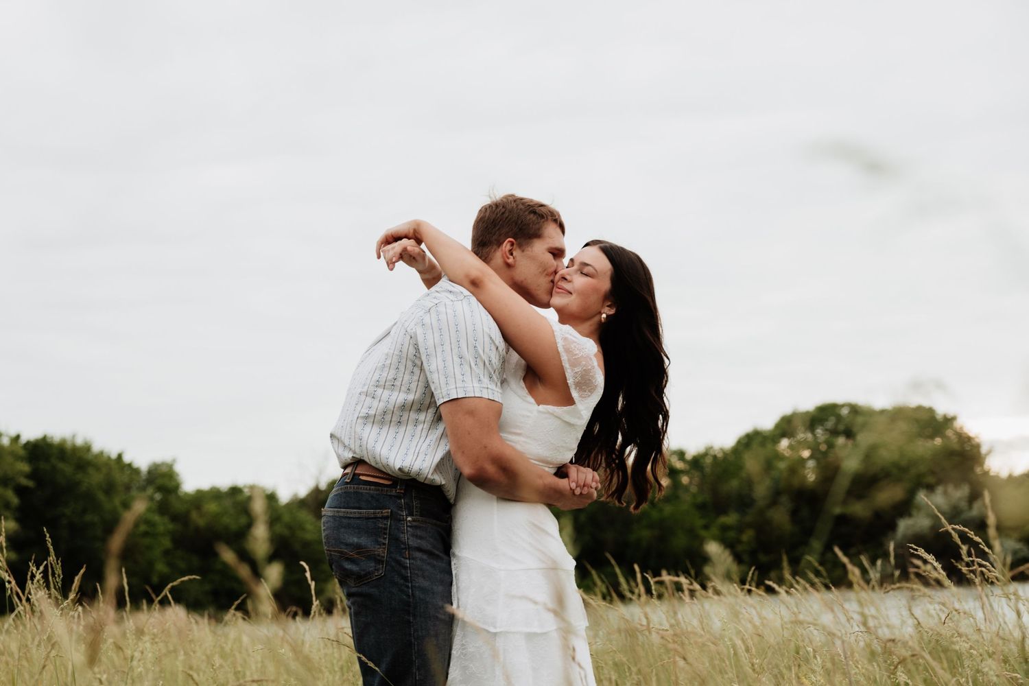 Couple sharing a romantic kiss in a golden grass field with lush green trees and overcast sky backdrop.