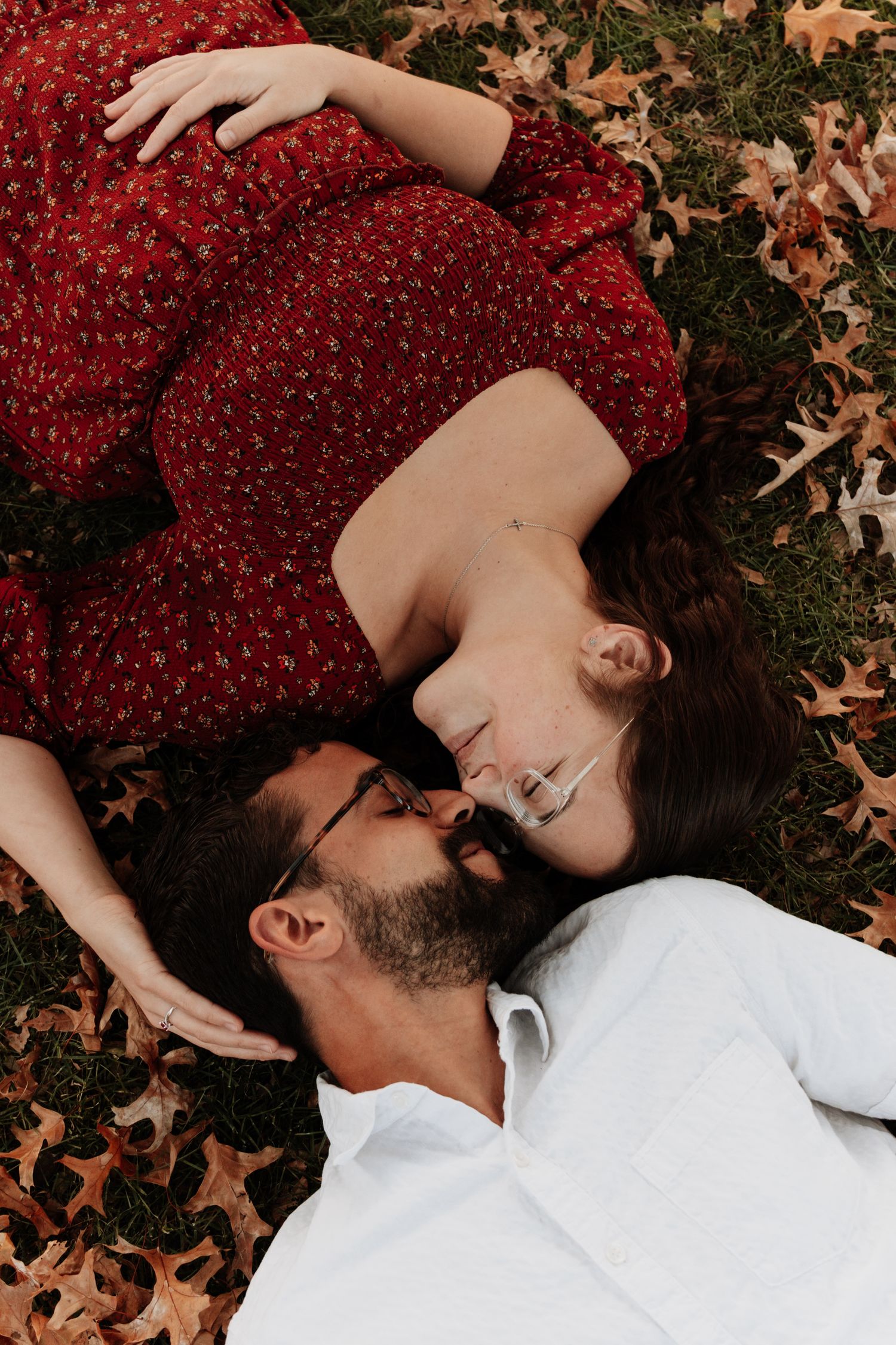 A couple lies on autumn leaves, with one wearing a red dress and the other in white.